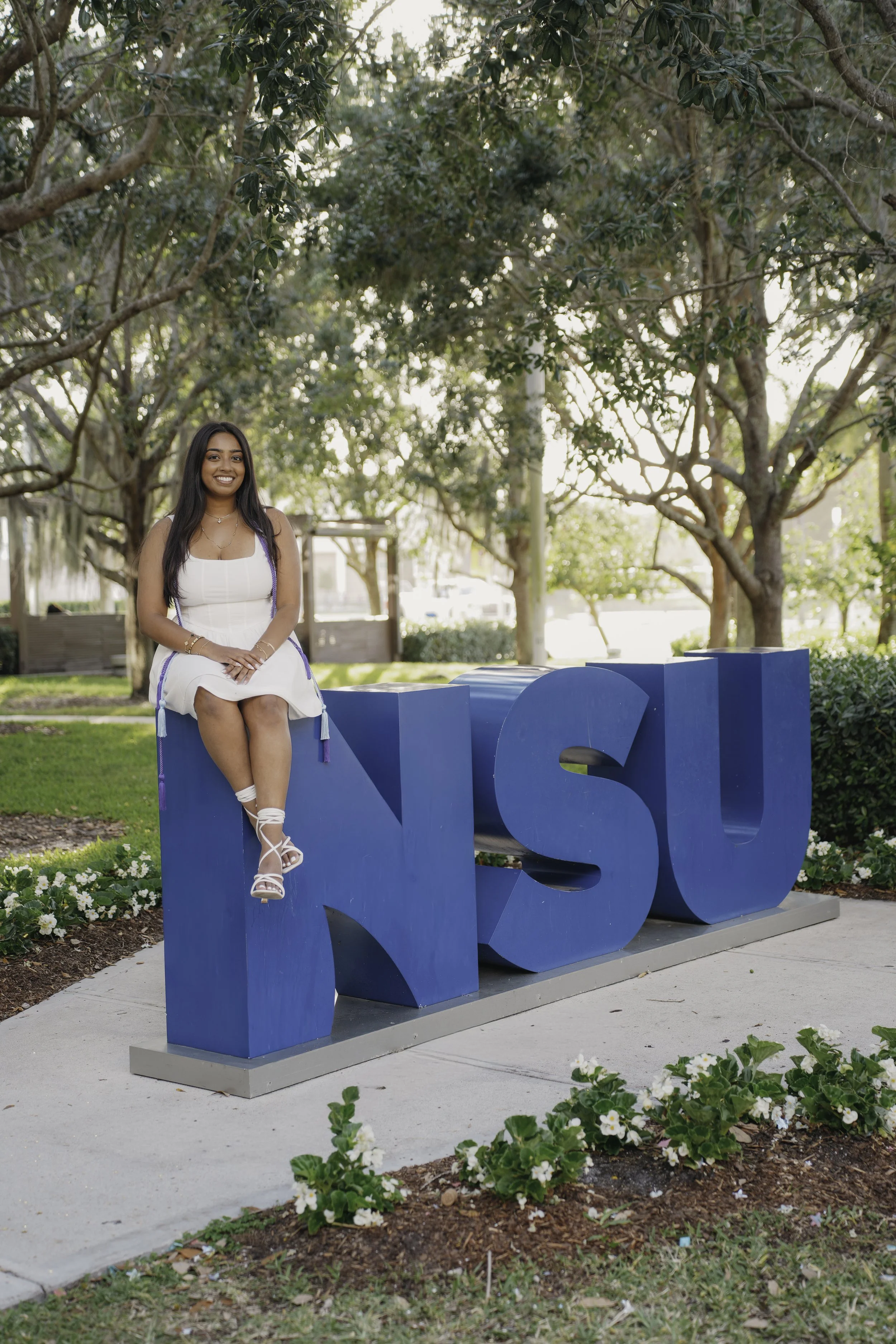 A young woman sitting on a large blue 'N' with the university letters in an outdoor park, smiling, wearing a white dress and sandals, surrounded by trees and flowers.