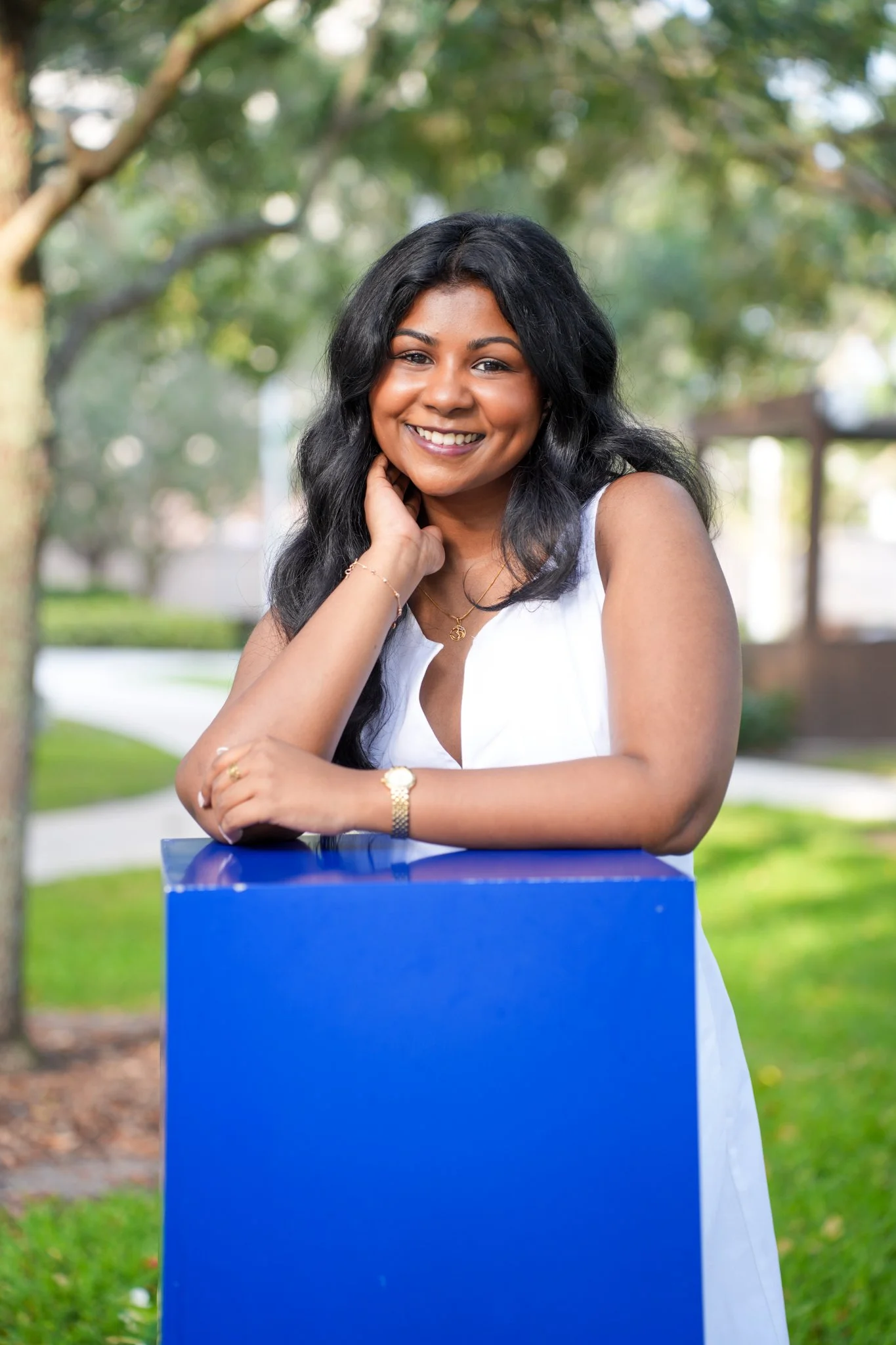 A smiling woman with black hair, wearing a white sleeveless dress, standing outdoors next to a blue rectangular object, with trees and greenery in the background.