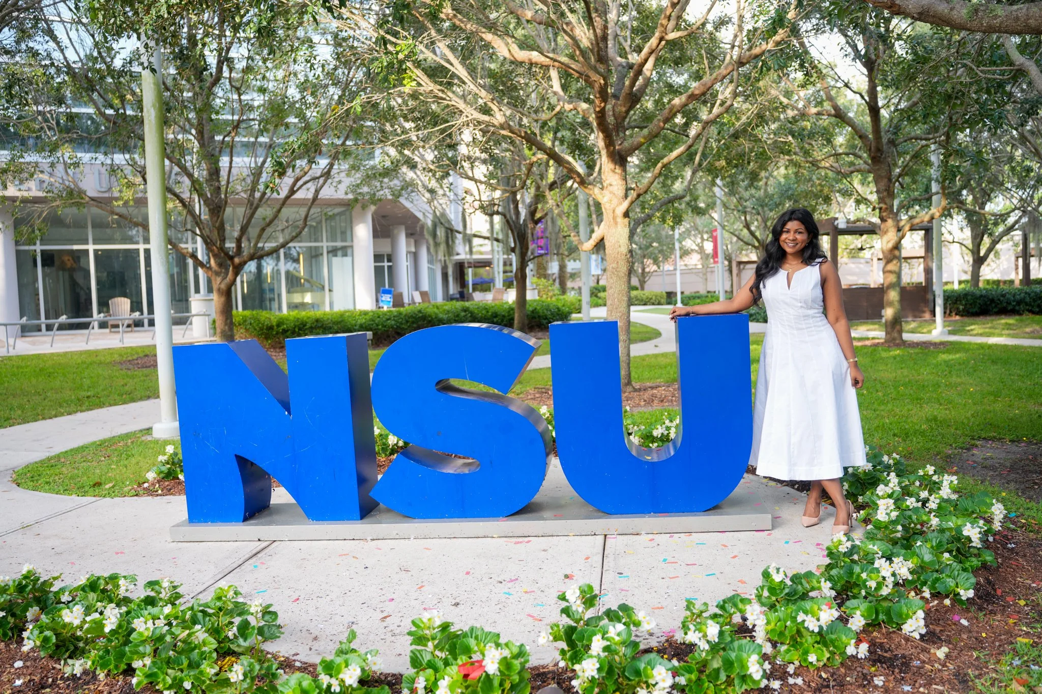 A woman in a white dress standing outdoors next to large blue letters spelling 'NSU' on a campus green with trees and modern buildings in the background.