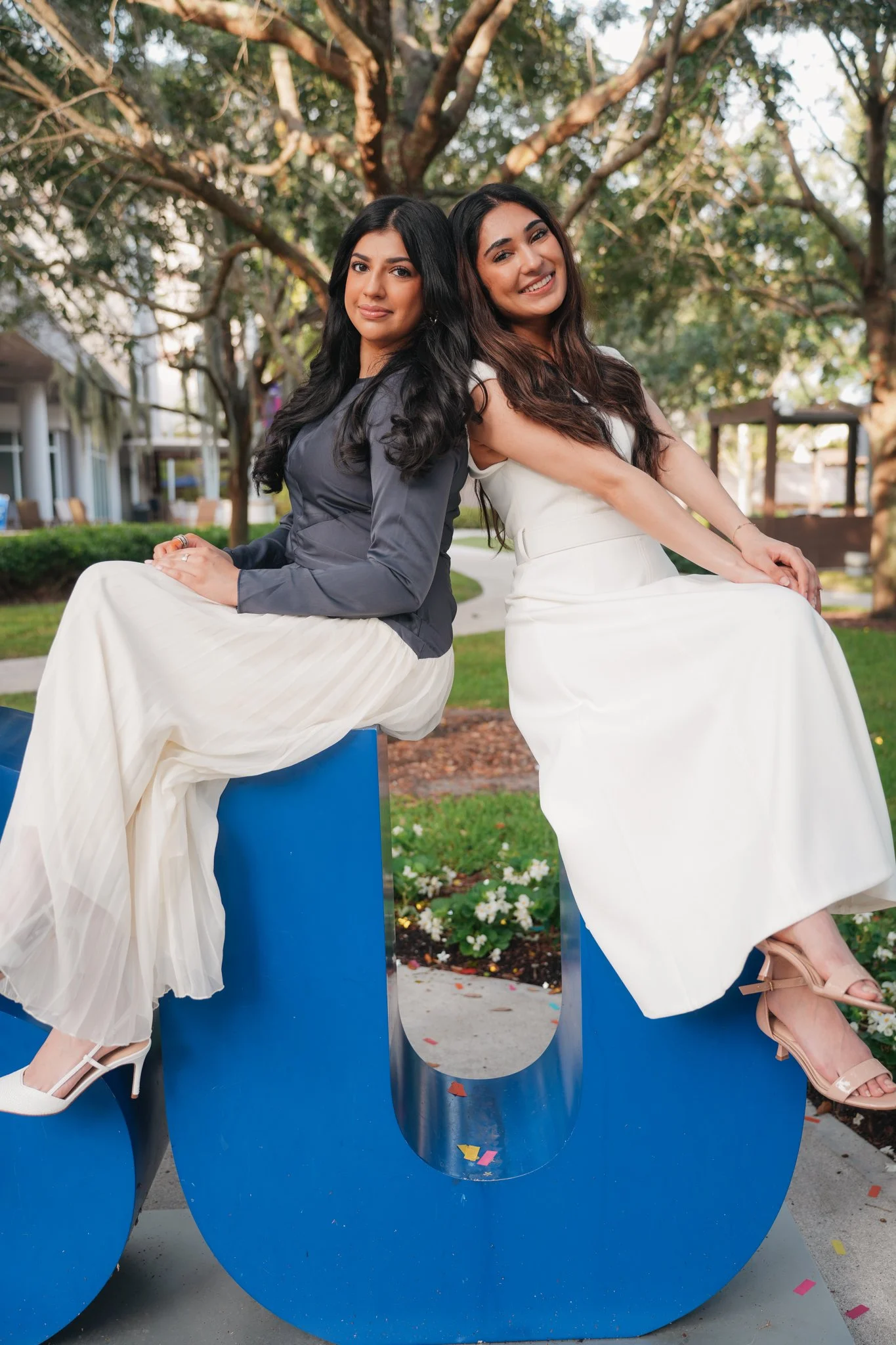 Two women sitting back-to-back on a large blue U-shaped sculpture outdoors, smiling; one wearing a white dress and the other a dark gray top and light-colored skirt, surrounded by trees and greenery.