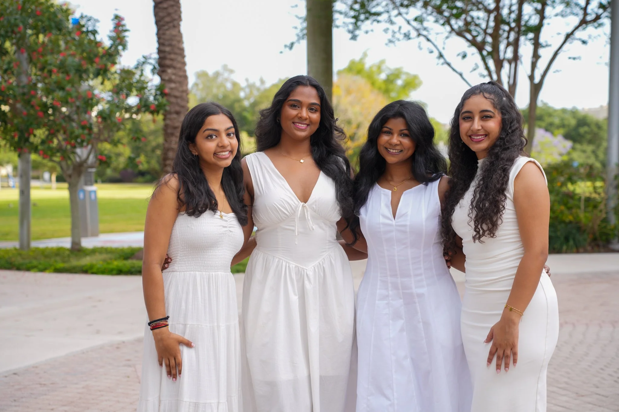 Four women standing outdoors in a park, smiling and dressed in white dresses.