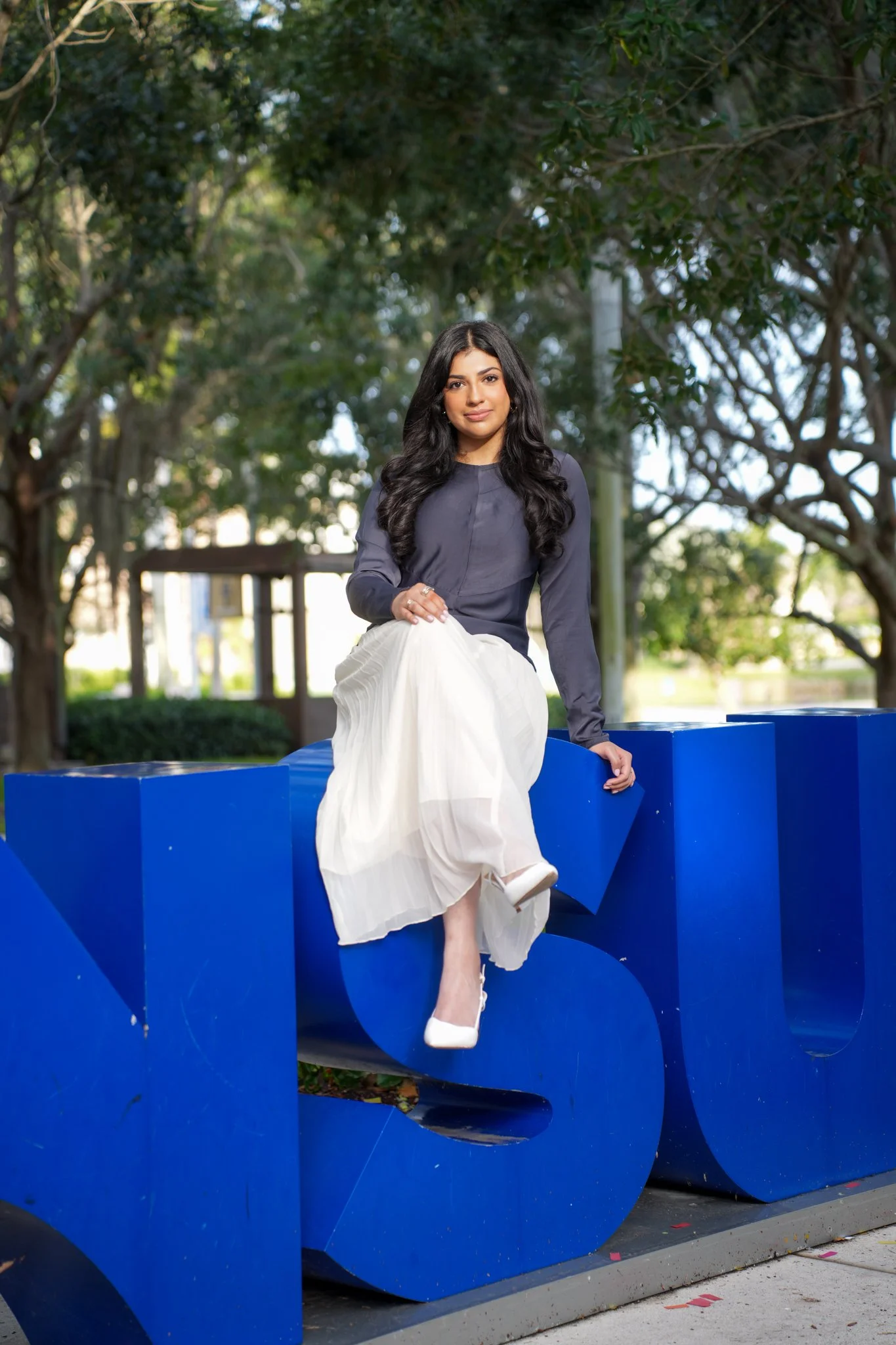 A woman in a navy blue long sleeve top and white pleated skirt sitting on a large blue 'S' sculpture outdoors surrounded by trees.