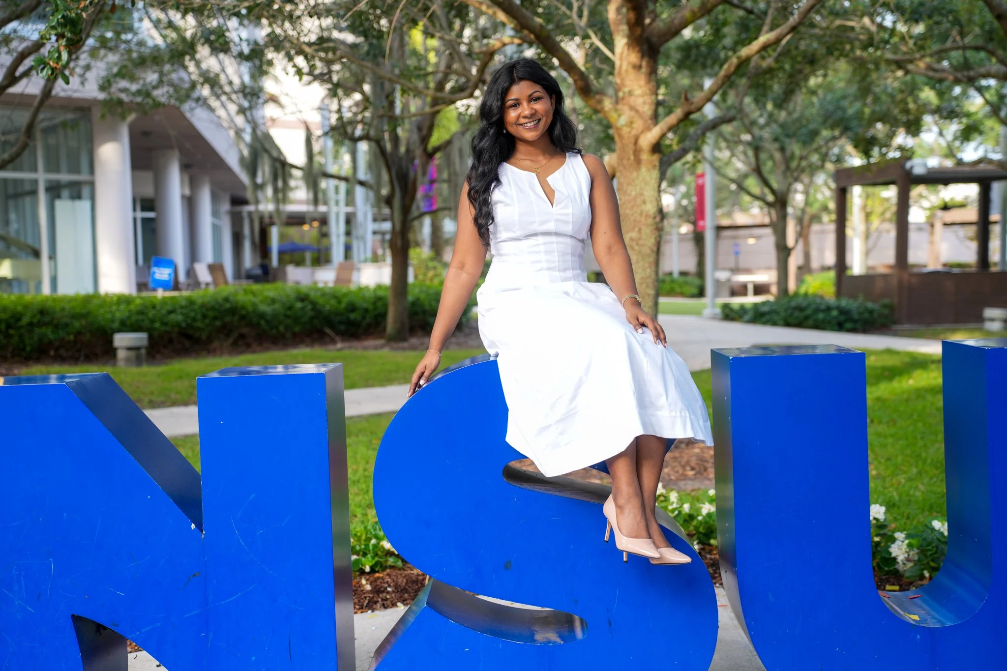 A woman in a white dress sitting on a blue sculpture in an outdoor park with trees and modern buildings in the background.