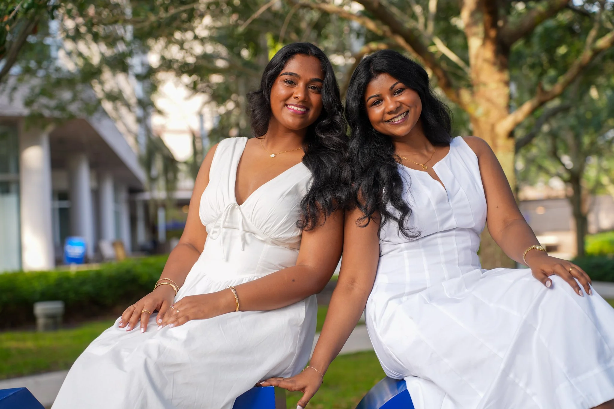 Two smiling women with long black hair wearing white dresses sitting outdoors on blue benches, with trees and a building in the background.