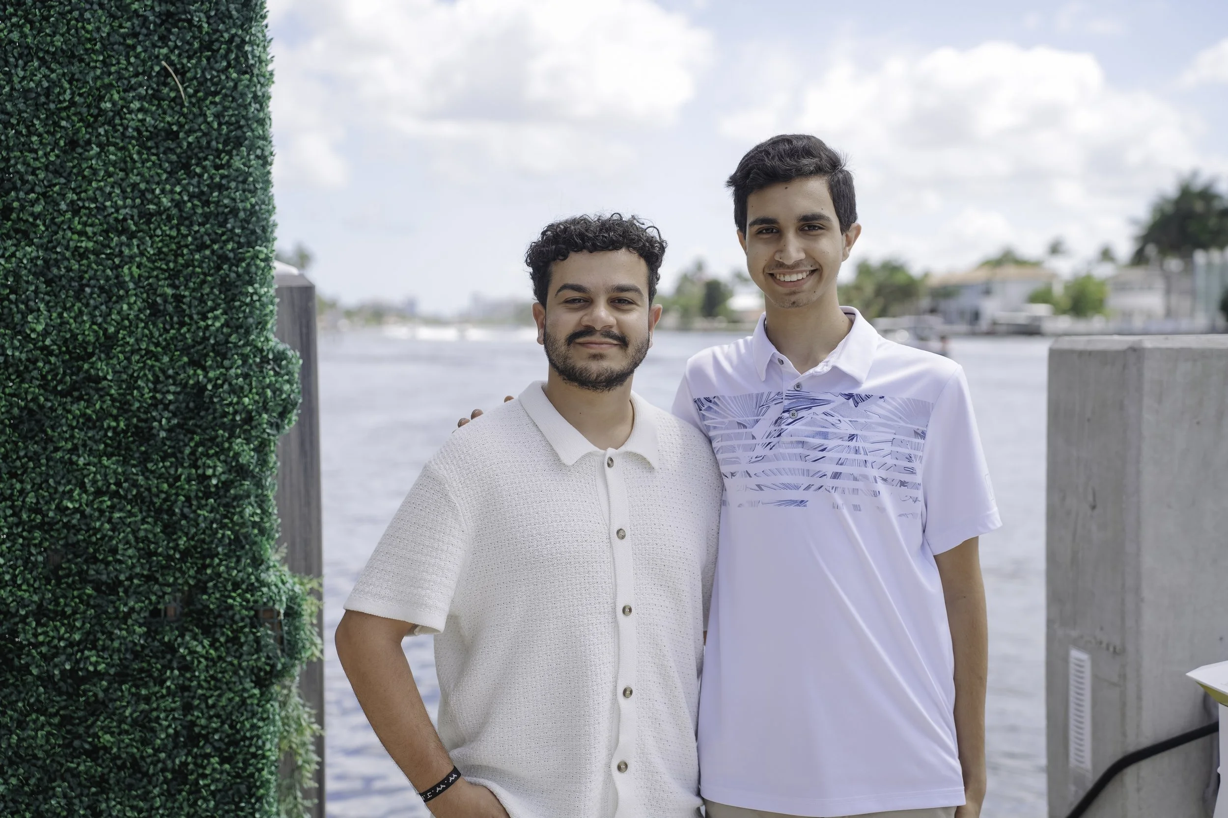Two young men smiling and standing beside each other near a body of water with houses and trees in the background, one wearing a white polo shirt and the other a white shirt with a blue pattern.