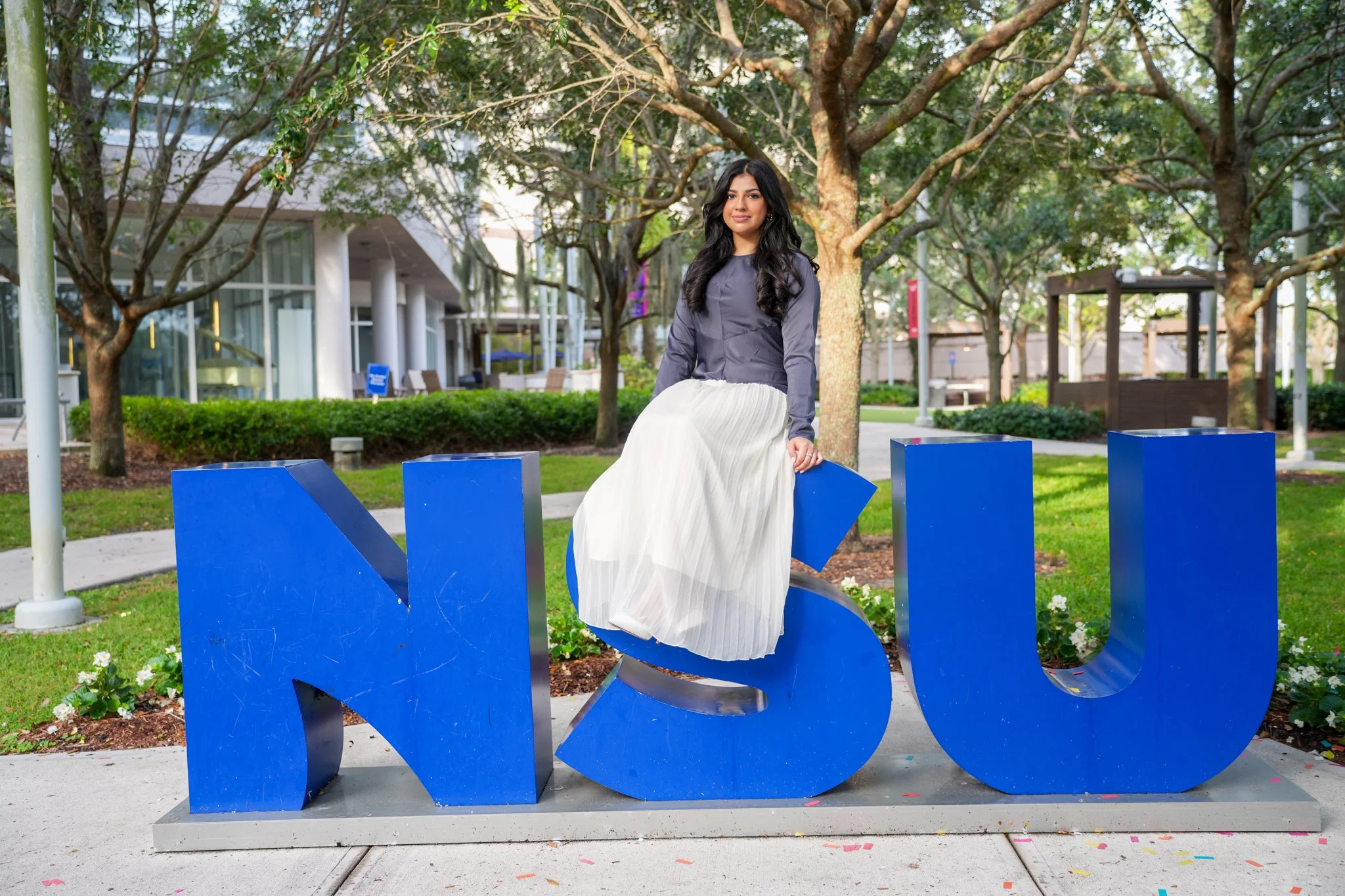 A young woman with long dark hair wearing a gray top and a pleated white skirt sitting on a large blue 'U' letter of the 'NOU' sign outdoors, surrounded by trees and a modern building.