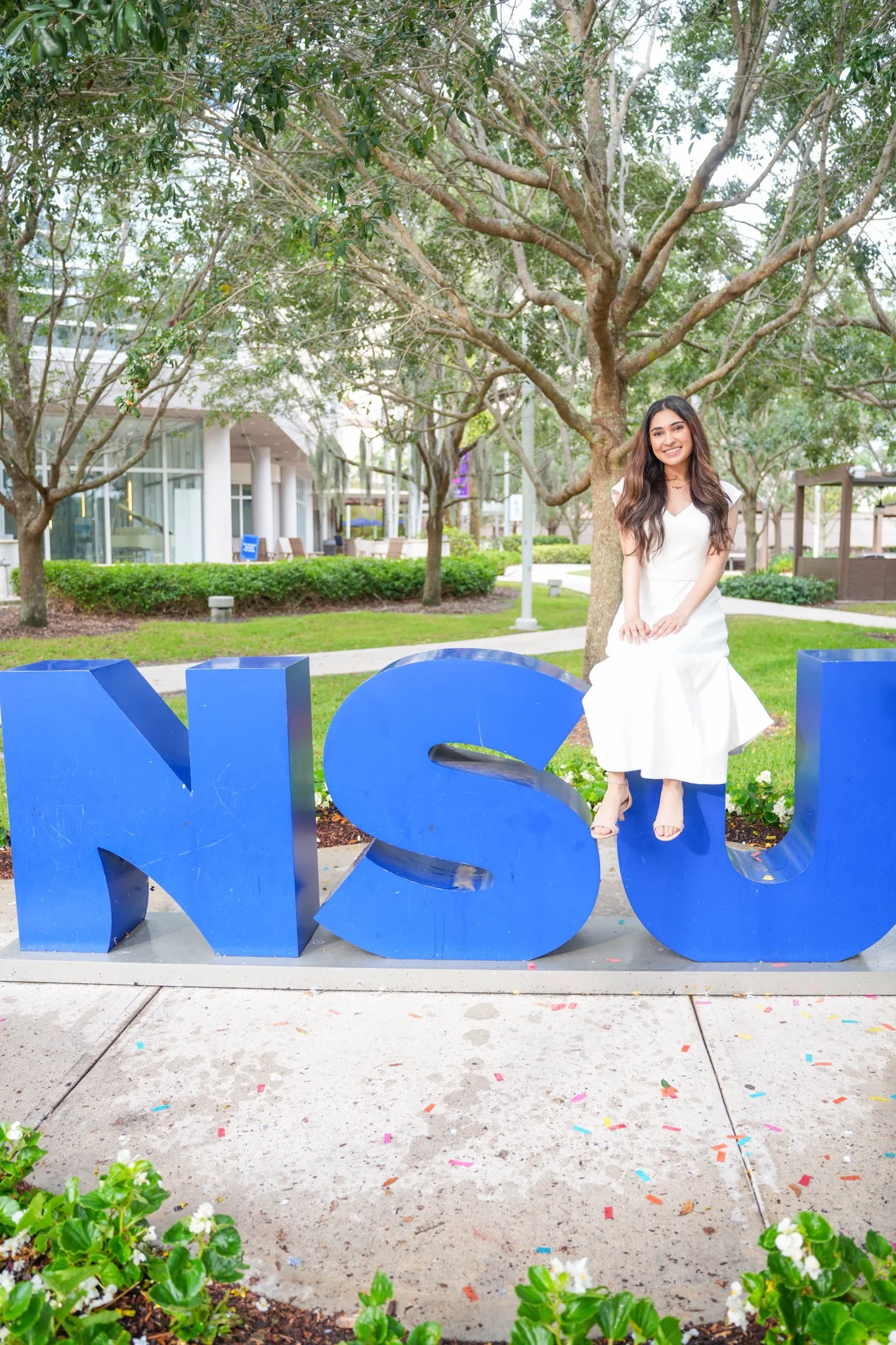 A young woman in a white dress sitting on large blue letters spelling 'NSU' outdoors in a park with trees and modern buildings in the background.