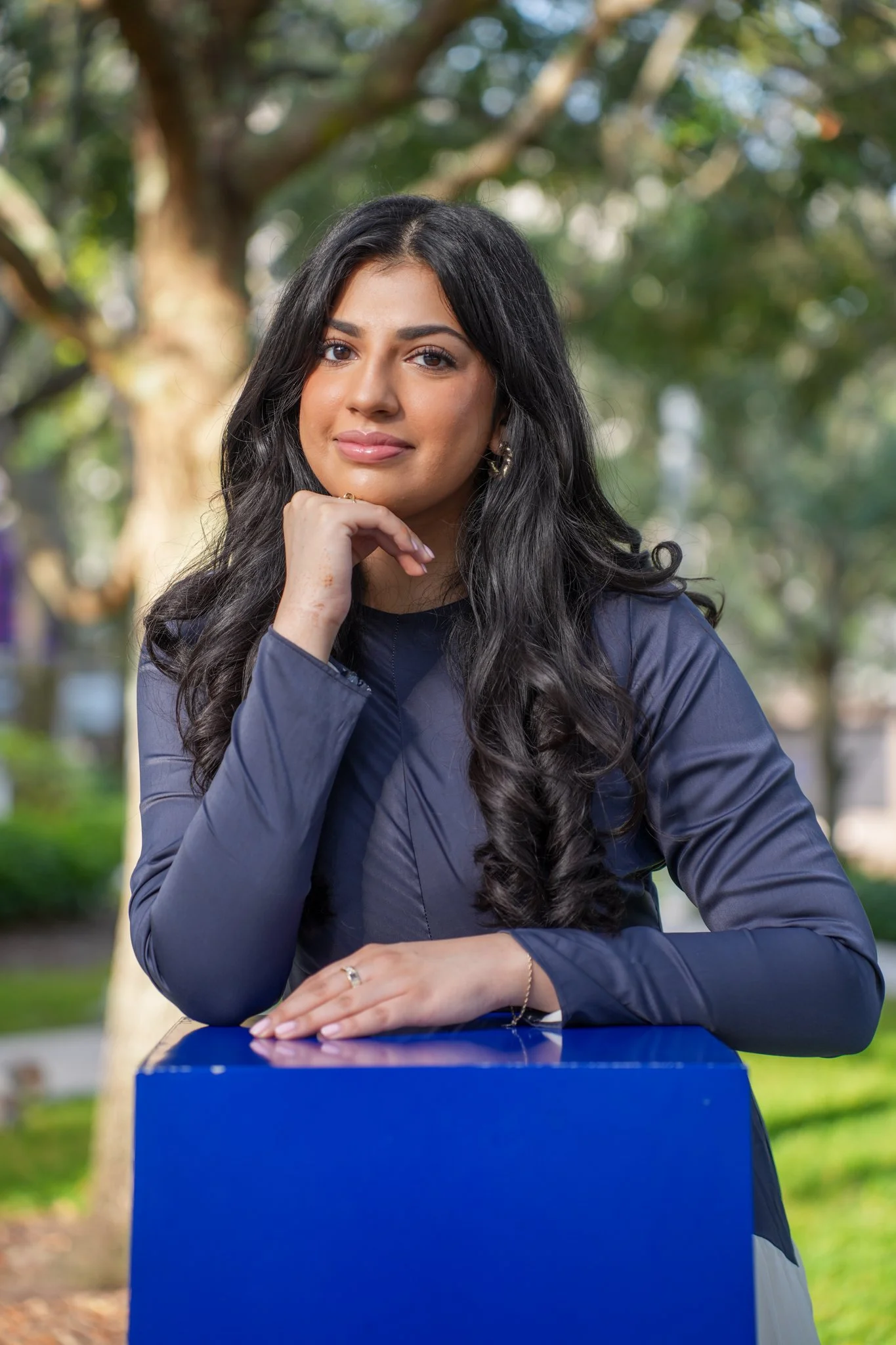A young woman with long black curly hair, wearing a dark gray jacket, sits outdoors with a background of trees and greenery, resting her chin on her hand and looking at the camera.