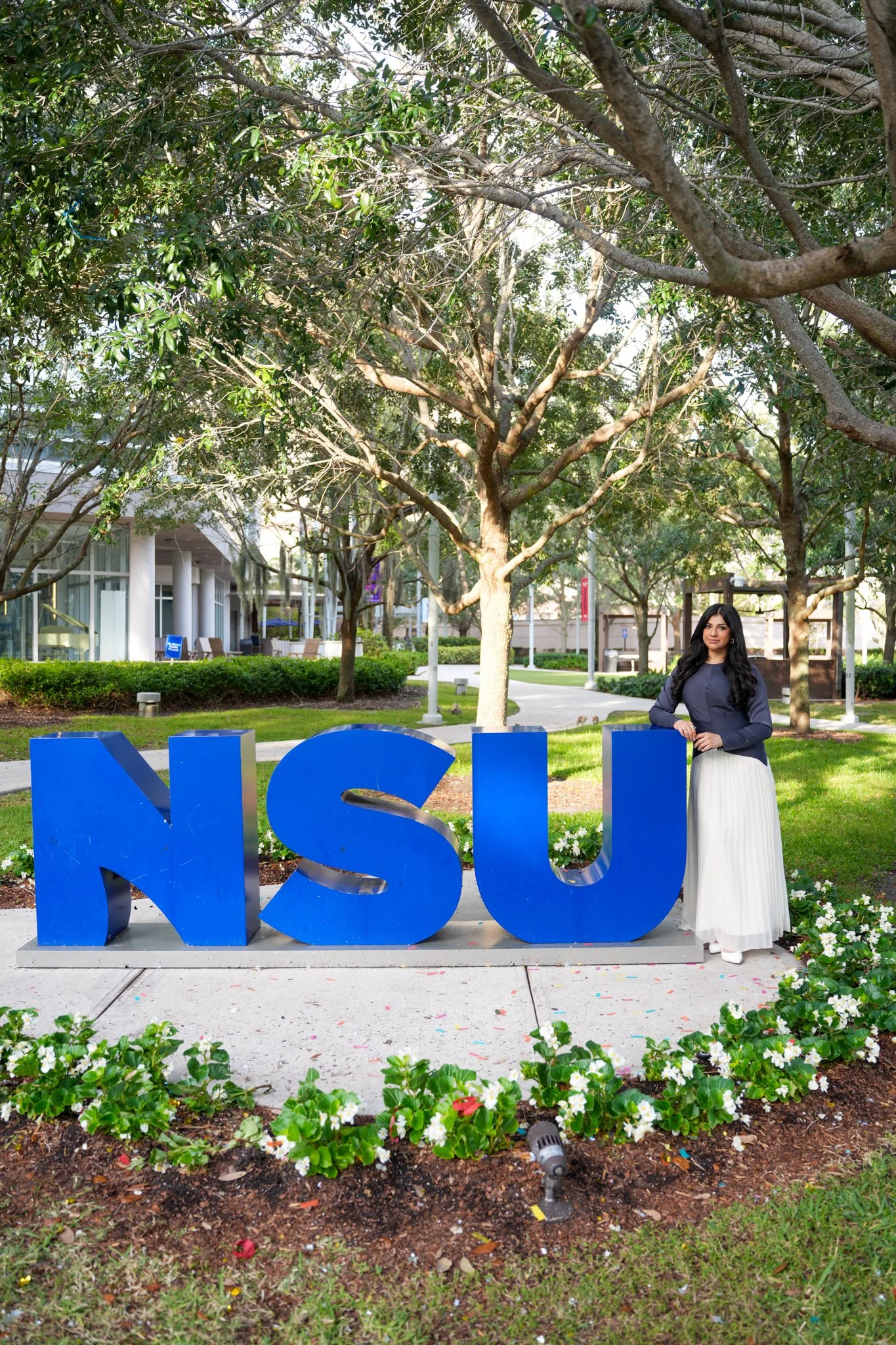 A woman stands beside large blue letters spelling 'NSU' in an outdoor campus setting with trees, grass, and a modern building in the background.