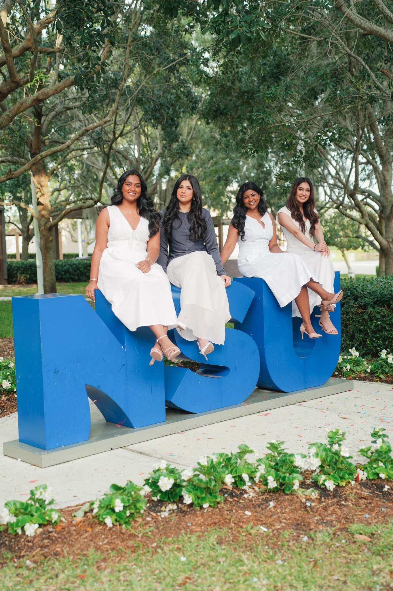Four women sitting on a large blue 'MSU' sign outdoors surrounded by trees and flowers.