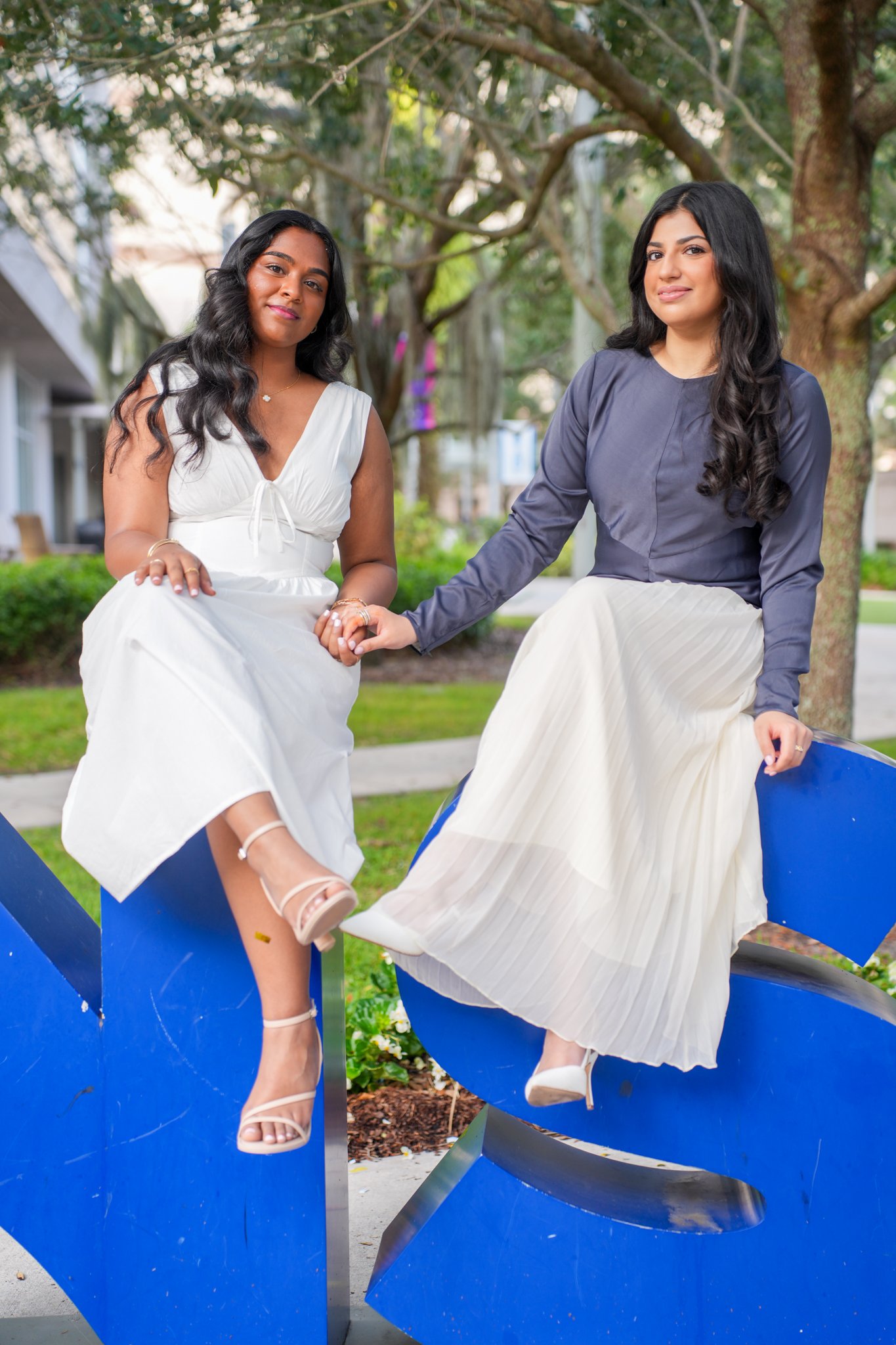 Two women sitting on a large blue abstract sculpture outdoors, holding hands and smiling. One woman wears a white dress and high heel sandals, the other wears a dark blouse and a cream pleated skirt.