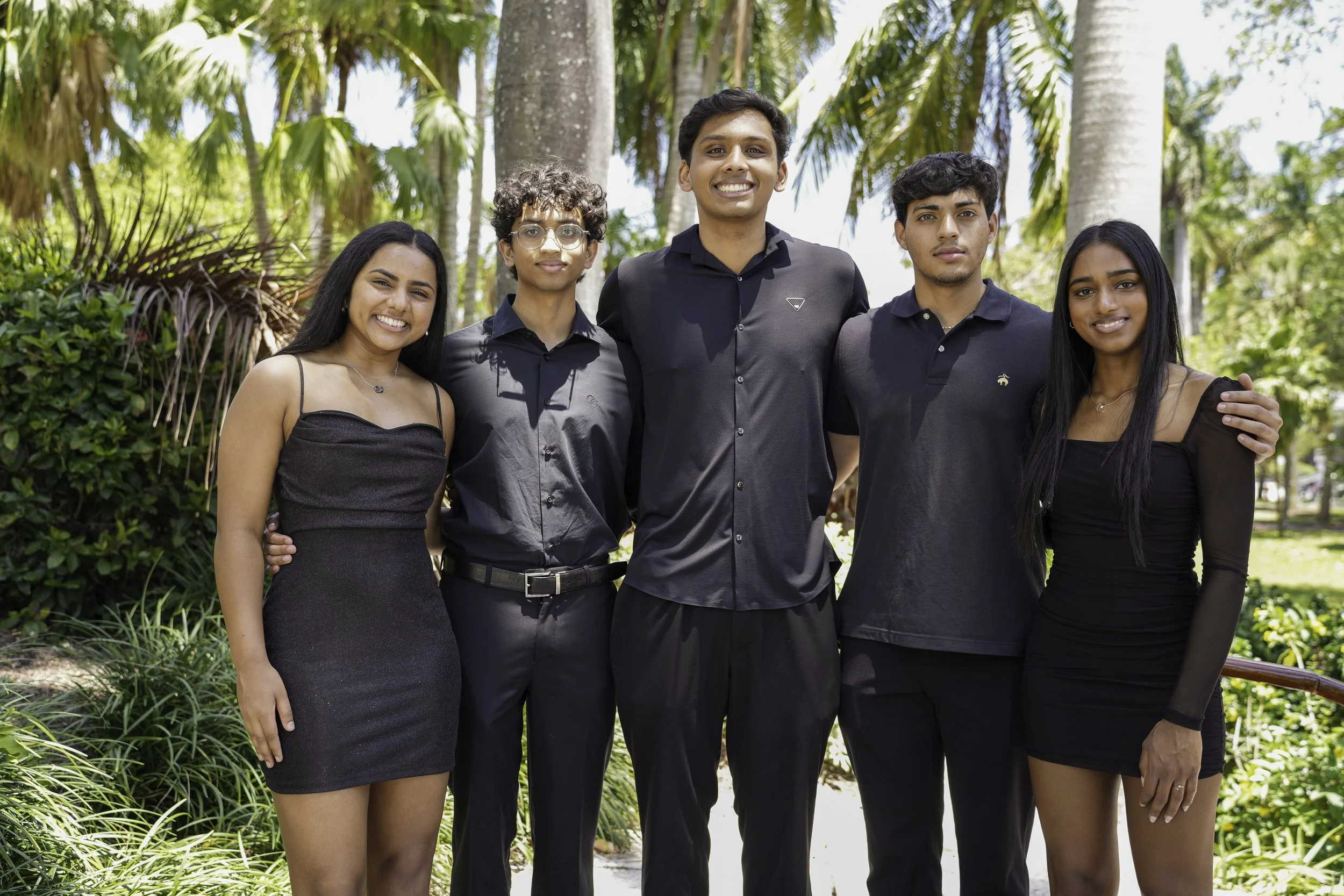 A group of five young people standing outdoors in a tropical park, dressed in black clothing, smiling and posing for a photo.