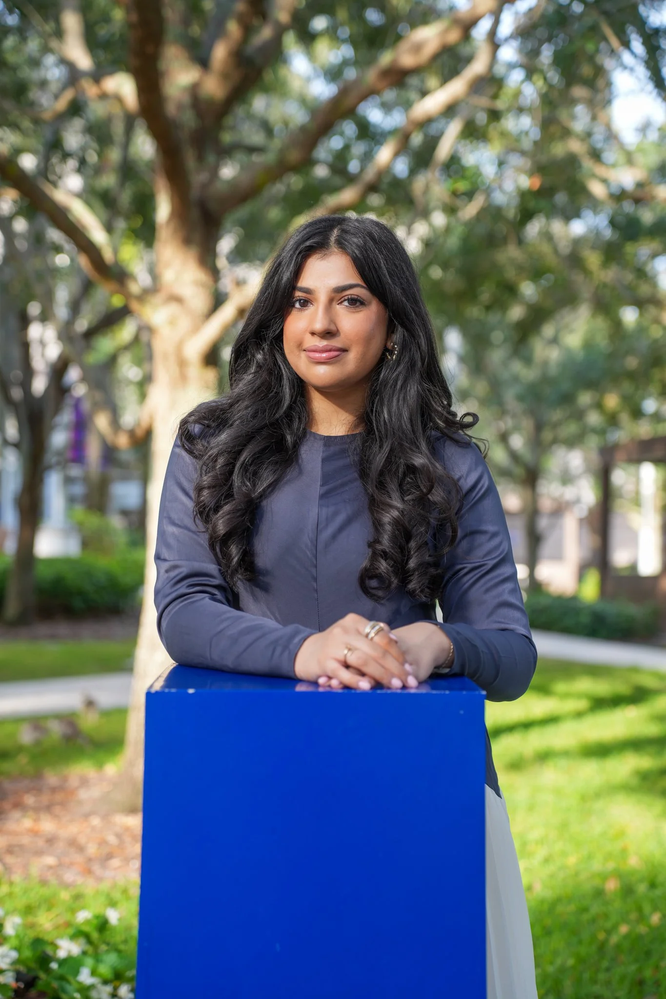 A woman with long black hair, wearing a dark long-sleeved shirt, stands outdoors with trees in the background, leaning on a blue podium.