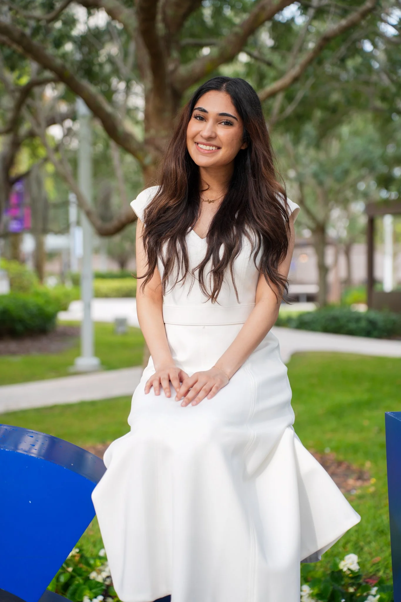 A young woman with long dark hair smiling and sitting outdoors in a white dress, with a leafy tree and park setting in the background.