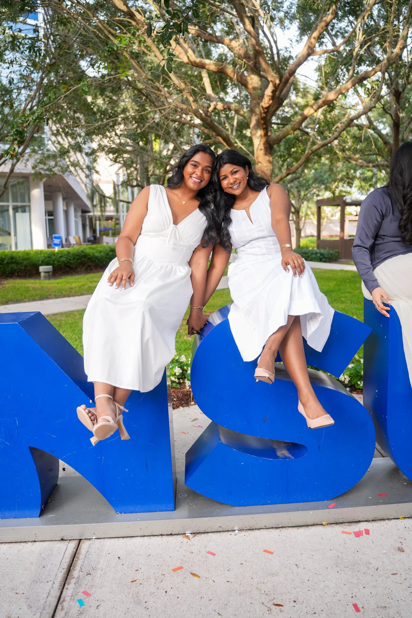 Two women sitting on large blue letters spelling 'MS' outdoors with trees and a building in the background.