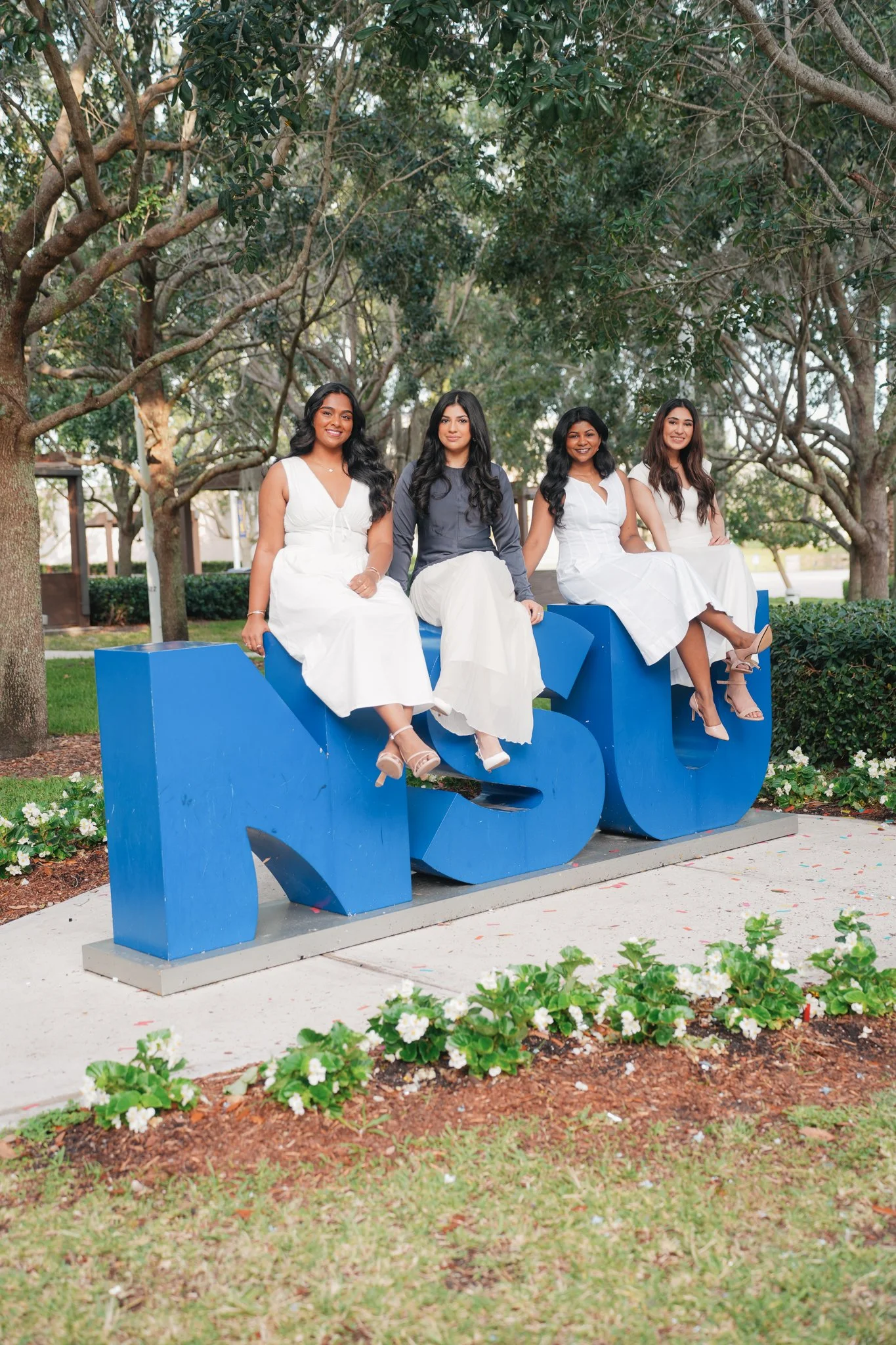 Four women in white dresses sitting on large blue letters spelling 'AUB' outdoors among trees and flowers.