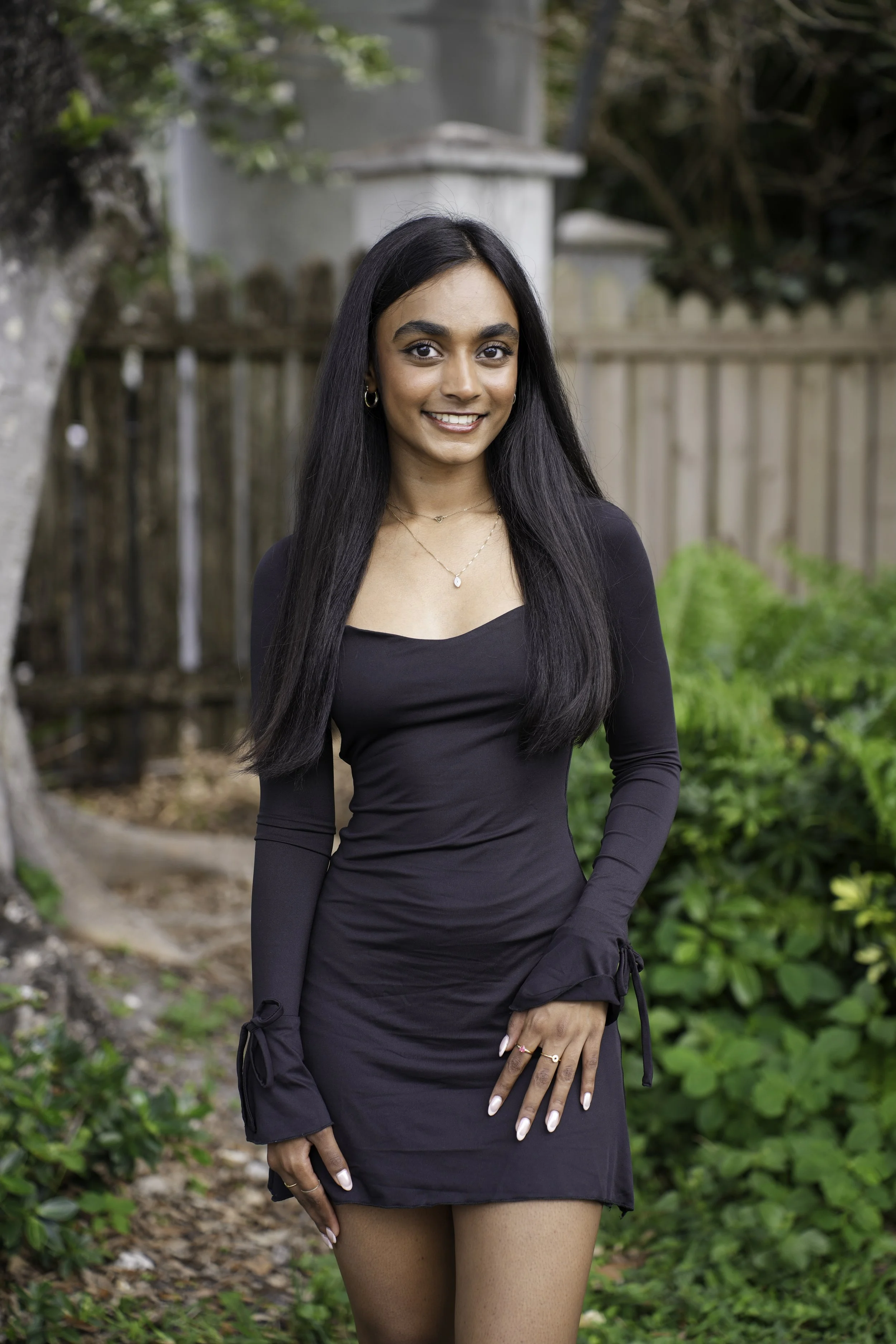 A young woman with long black hair, wearing a black dress with long sleeves and tied cuffs, standing outdoors in a garden with a wooden fence, green plants, and trees in the background.