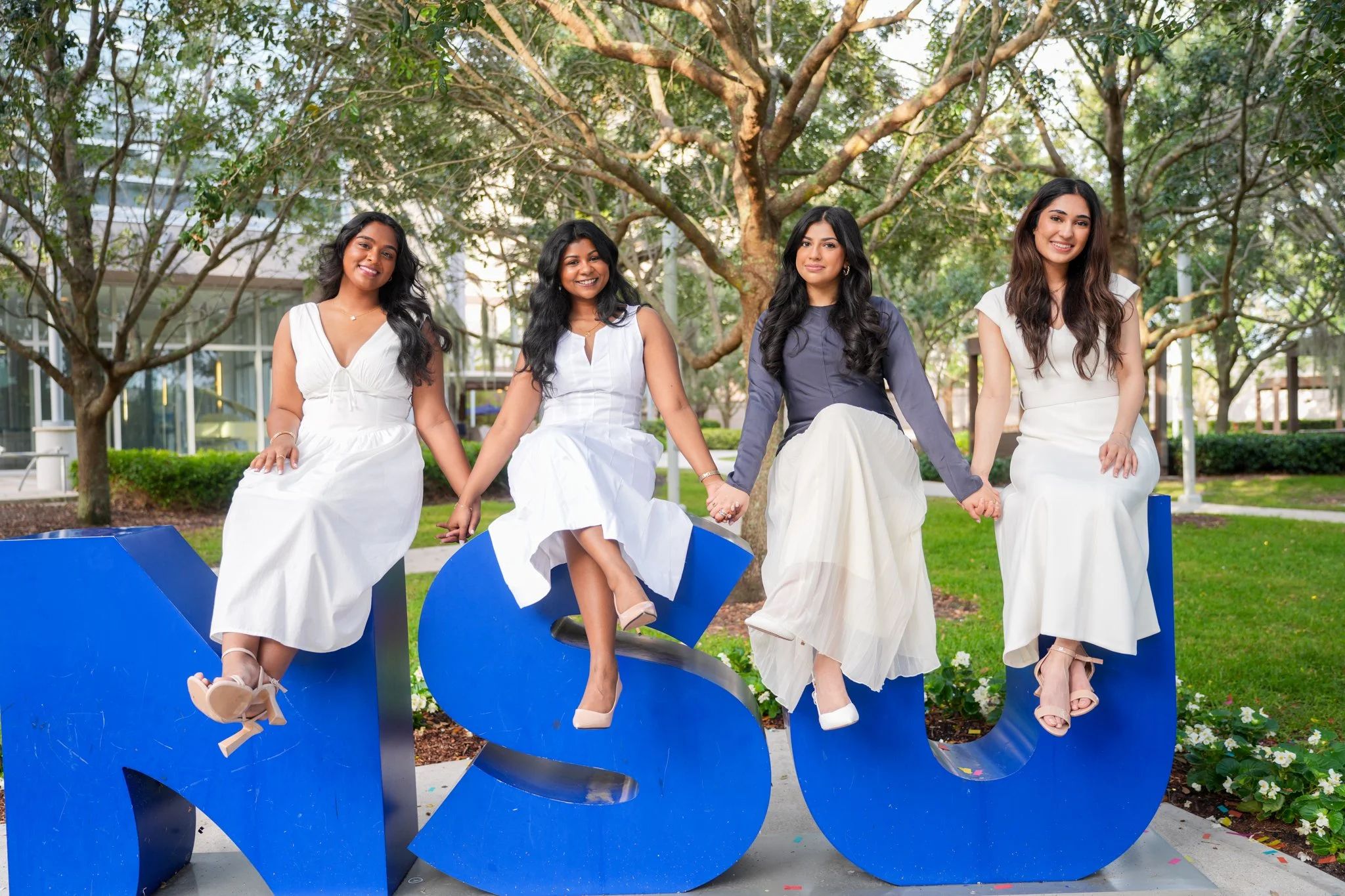 Four young women in white and navy dresses sitting on large blue letters spelling 'MSU' outdoors, smiling with trees and modern buildings in the background.