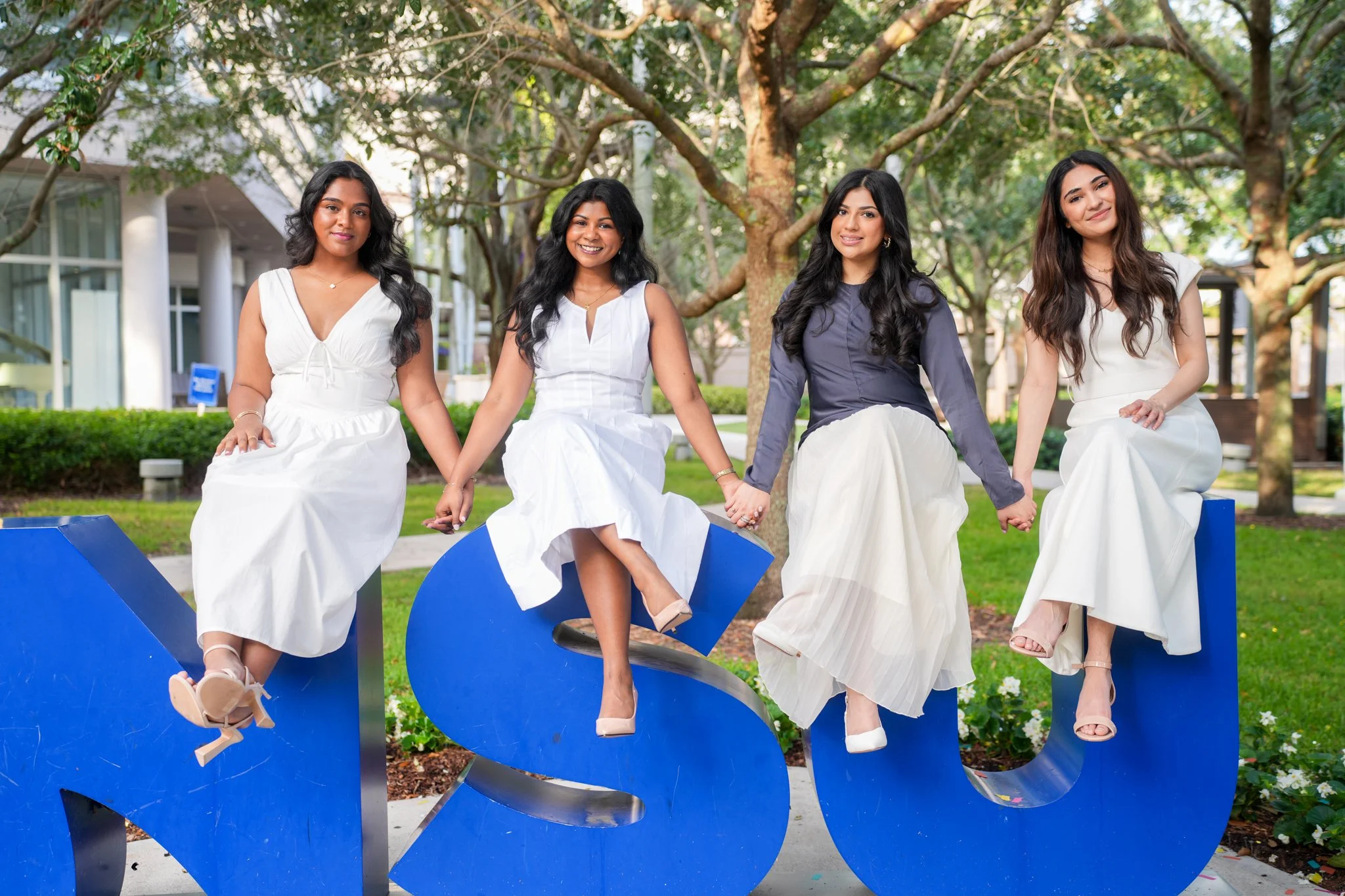 Four women dressed in white and dark clothing, sitting on large blue letters spelling 'MSU' outdoors in a park-like setting with trees and buildings in the background.