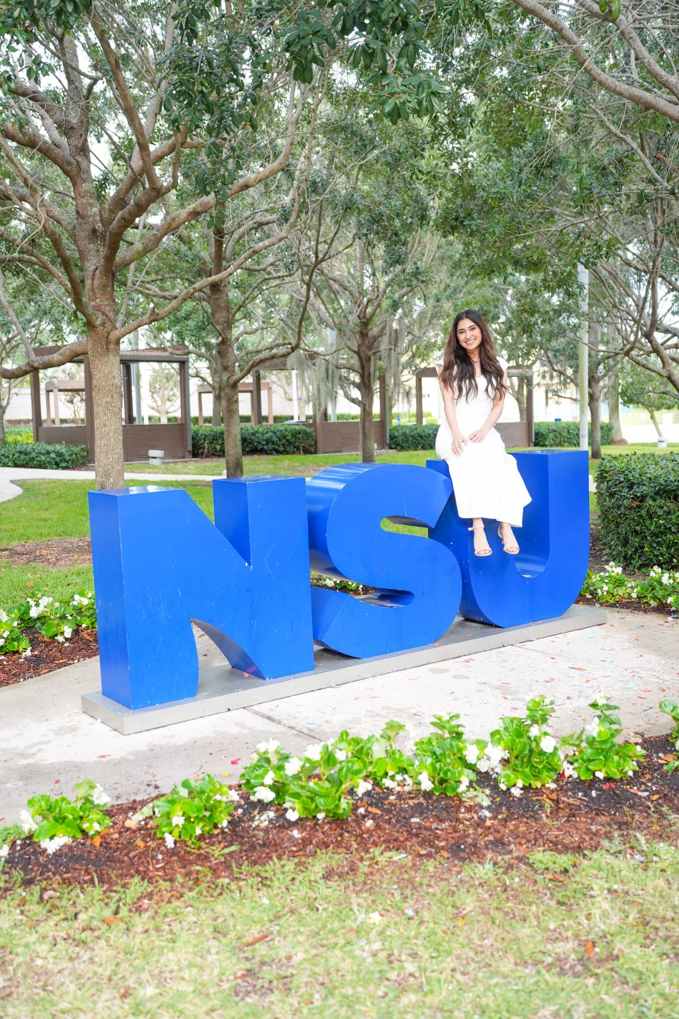 Young woman sitting on a large blue 'NSU' sculpture in a park with trees and greenery.