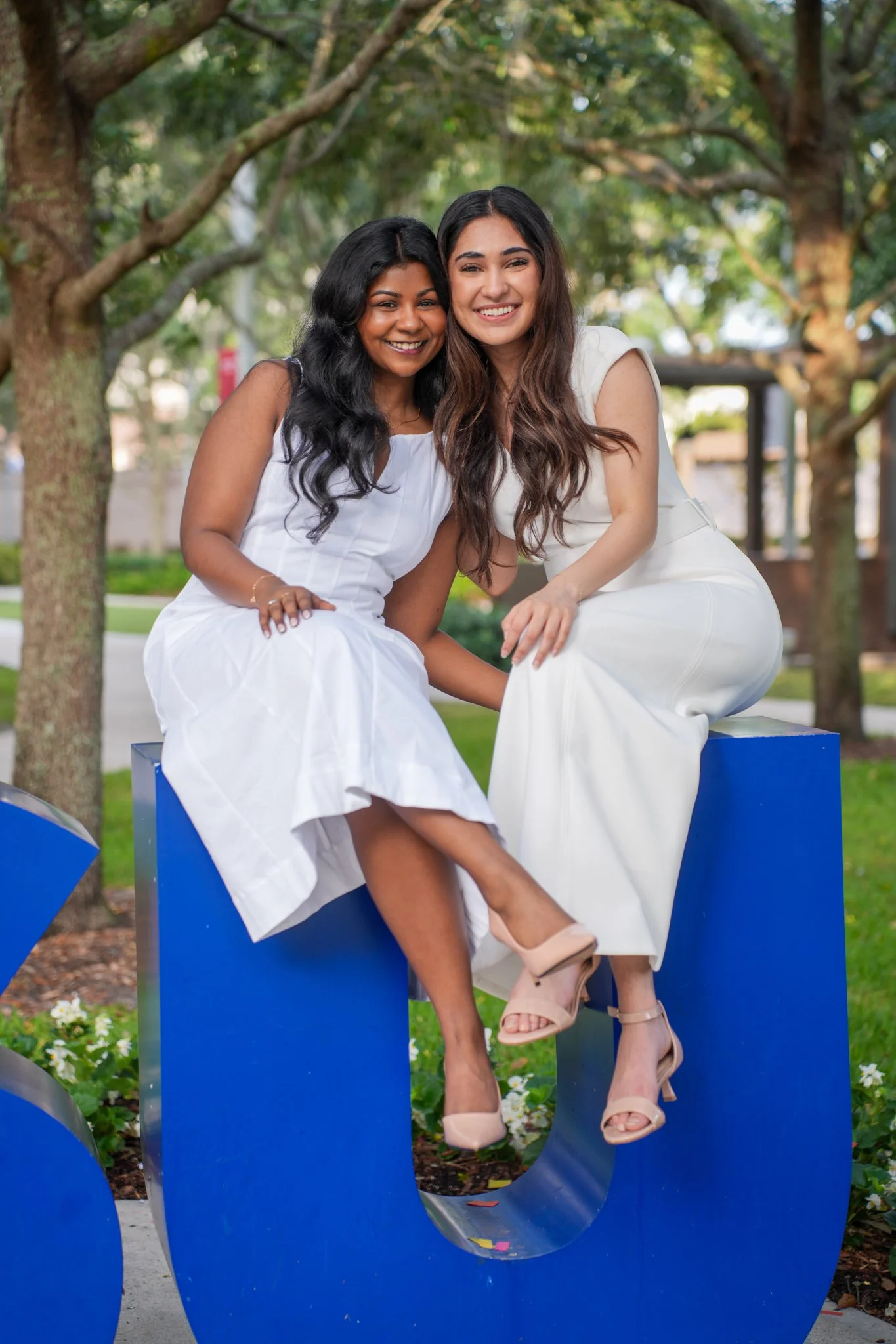 Two women sitting on a blue sculpture outdoors, smiling and embracing in a park with trees in the background.