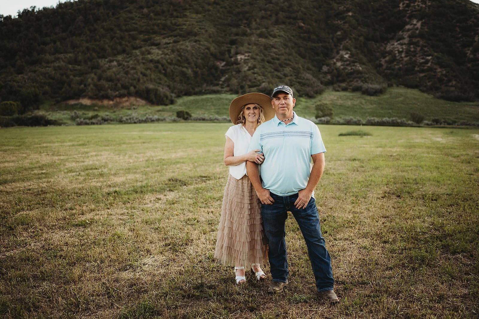 A couple standing arm-in-arm in an open grassy field with hills and mountains in the background, smiling at the camera.