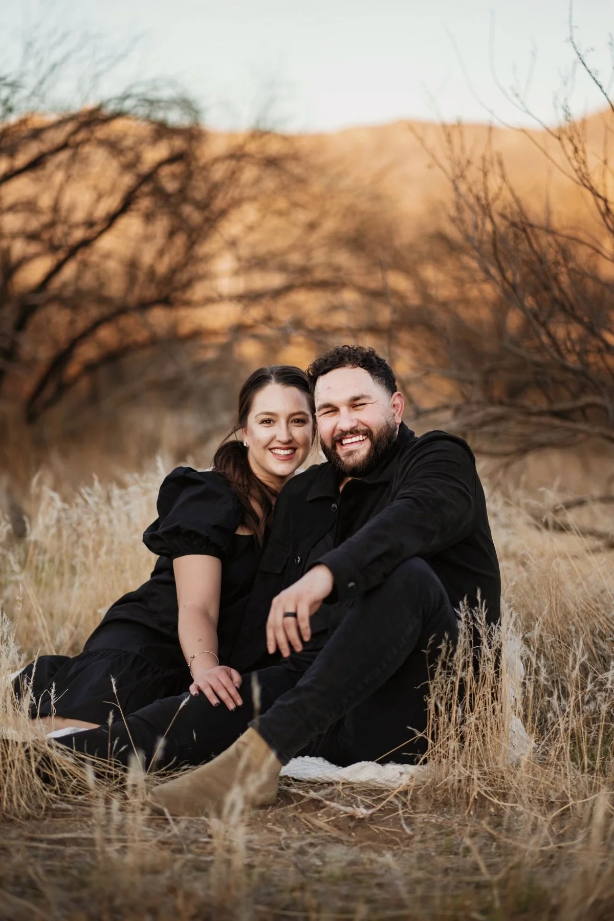 Nate & Sabrina Yanez. A happy couple sitting on the ground in a field with dried grass, smiling at the camera, with dry trees and hills in the background.
