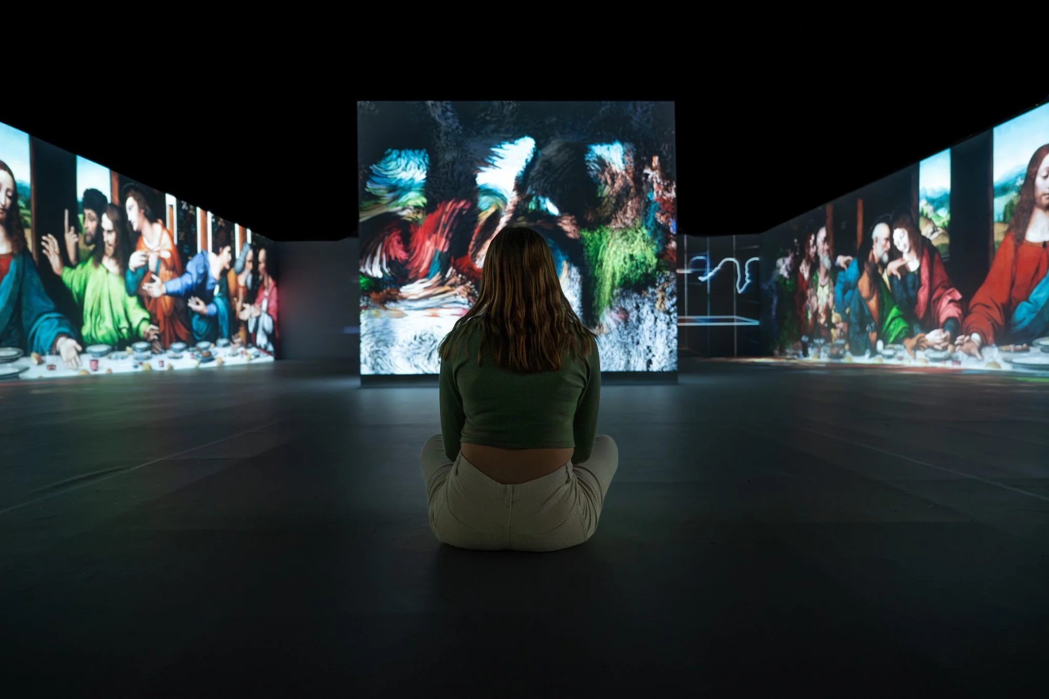 A woman sitting cross-legged in a dark art gallery, surrounded by large digital screens displaying classical paintings and abstract digital art.