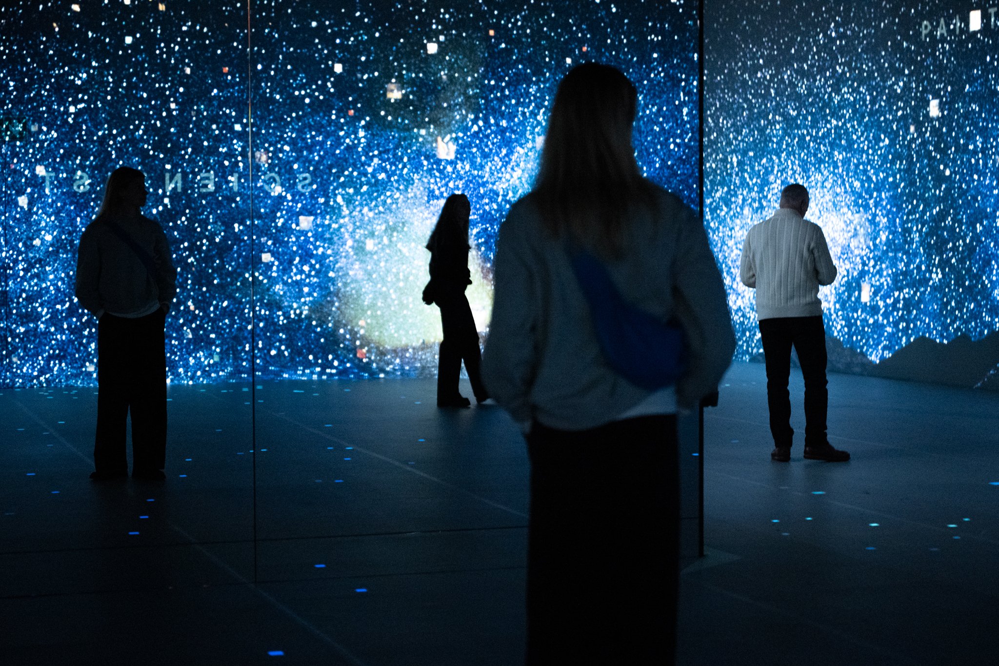 People observing an interactive digital art installation resembling a starry night sky with blue and white specks.