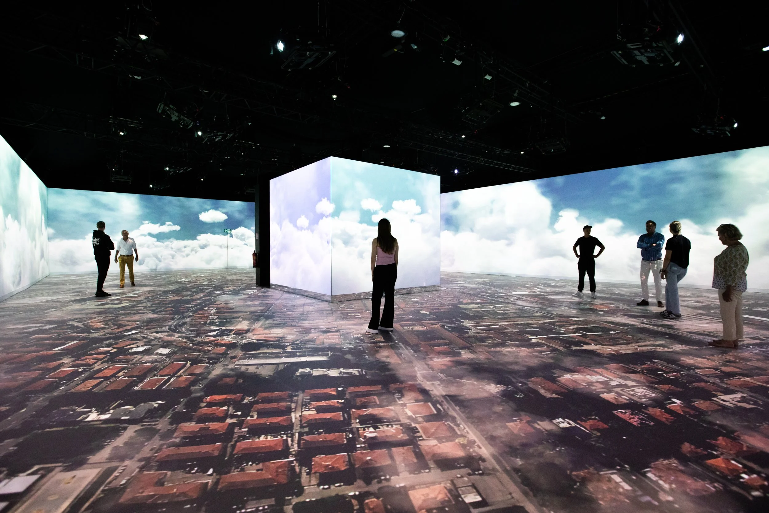 People observing an immersive digital art installation with a sky and cloud display on large screens surrounding a space with aerial map imagery on the floor.