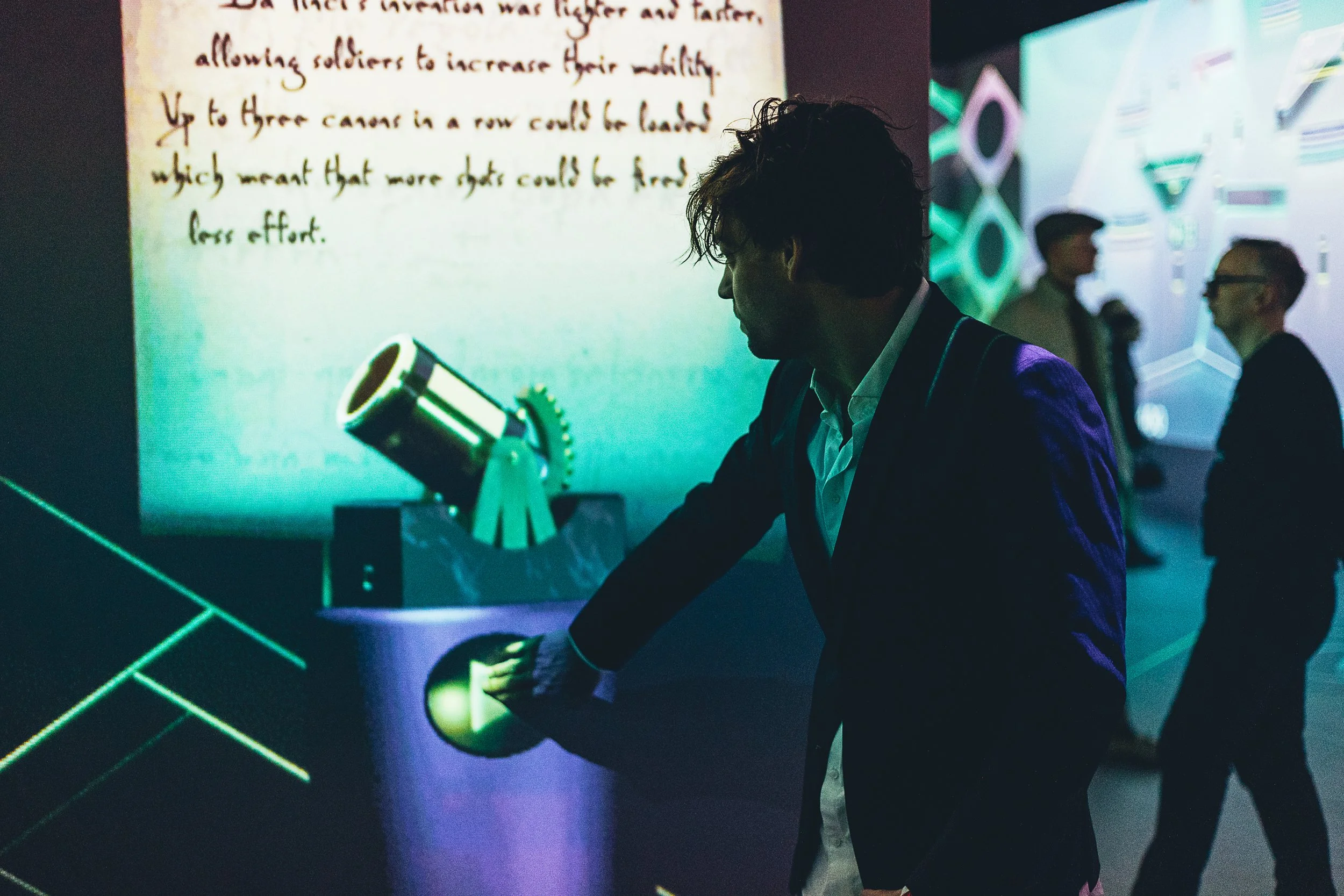 Person in dark suit and white shirt interacting with a device emitting green light, at a tech exhibit. In the background, a large screen displays handwritten text about a historical topic, and other people are talking near illuminated geometric wall patterns.