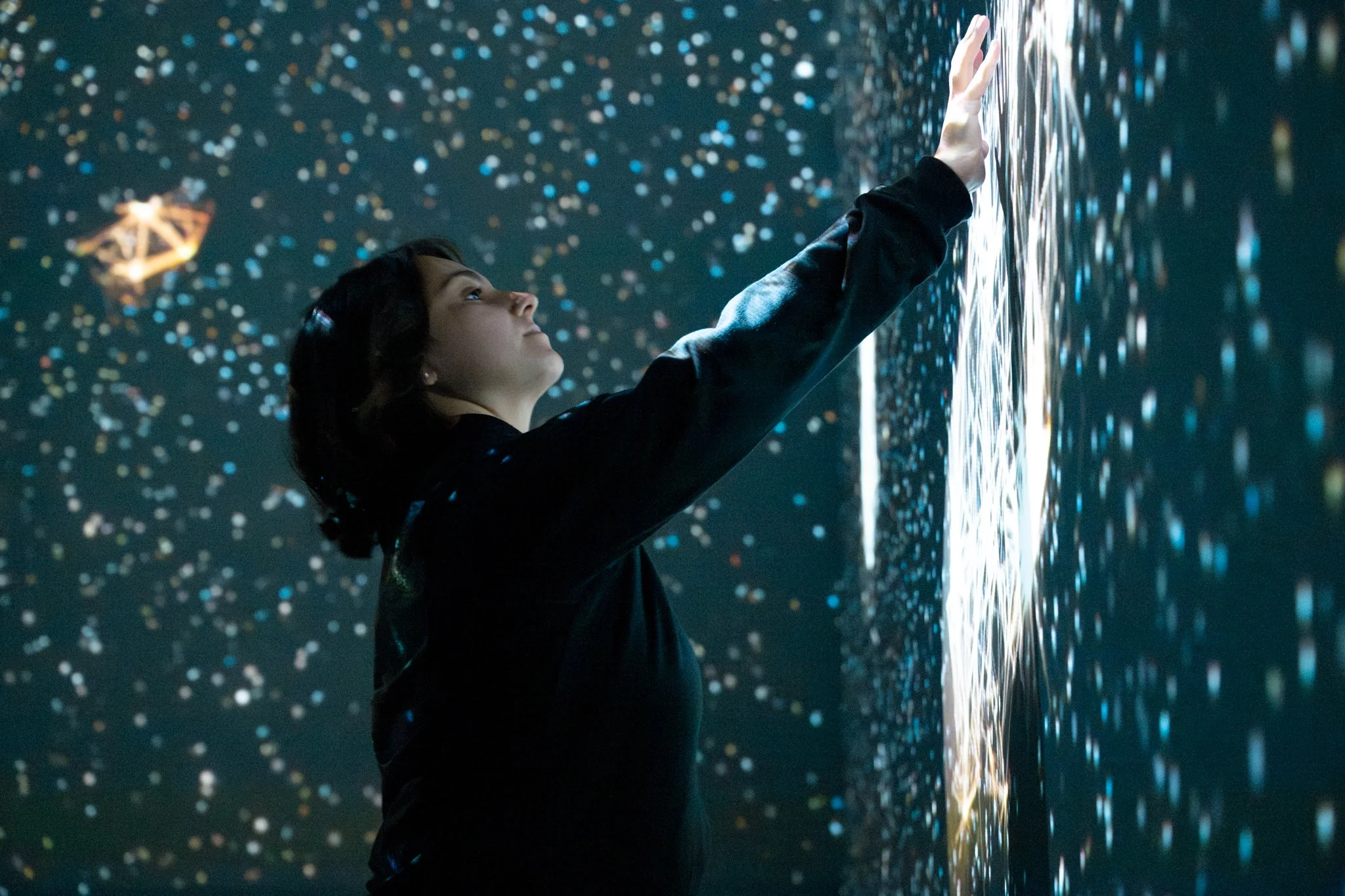 A woman touching a digital display wall with floating colorful particles around her.