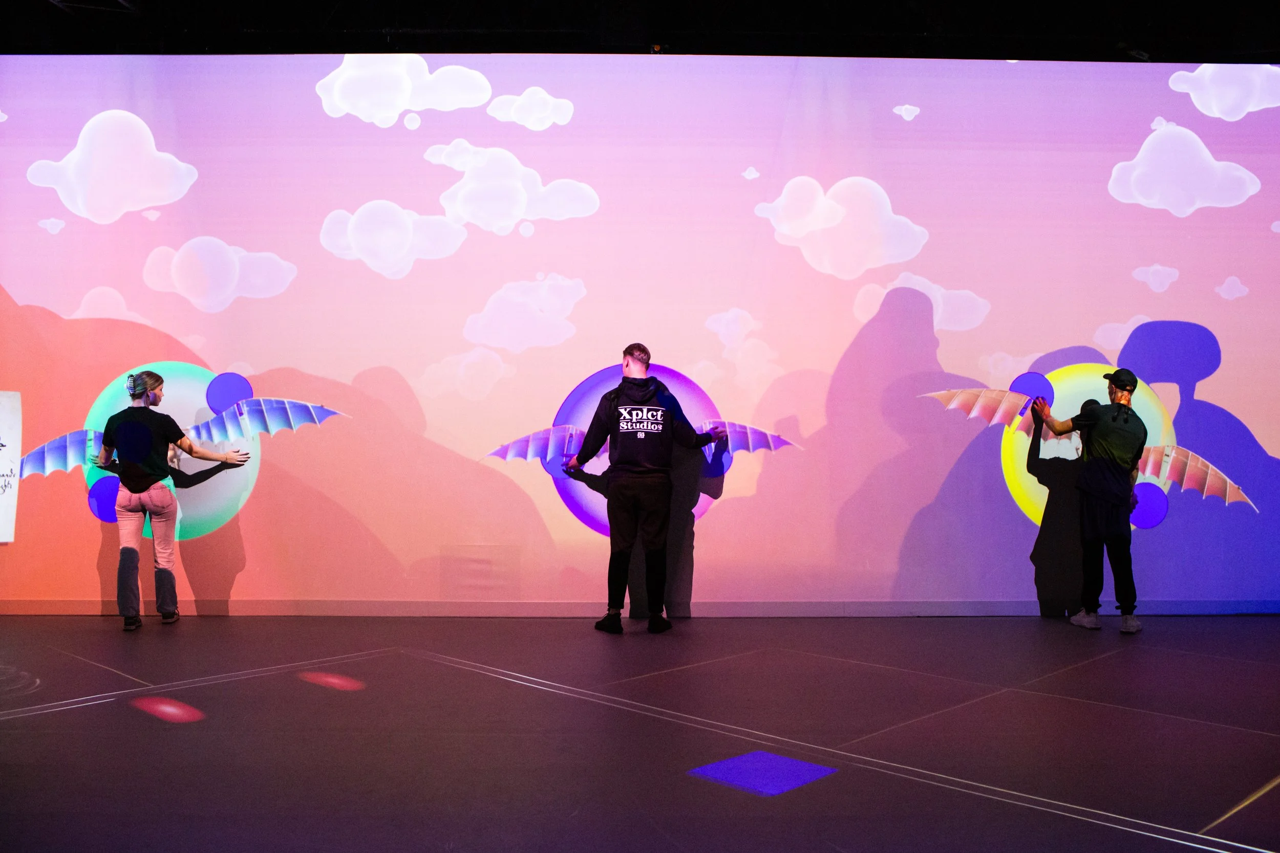 Three people are creating and adding digital illustrations of wings to a large projection screen. The background shows a pastel-colored sky with clouds.