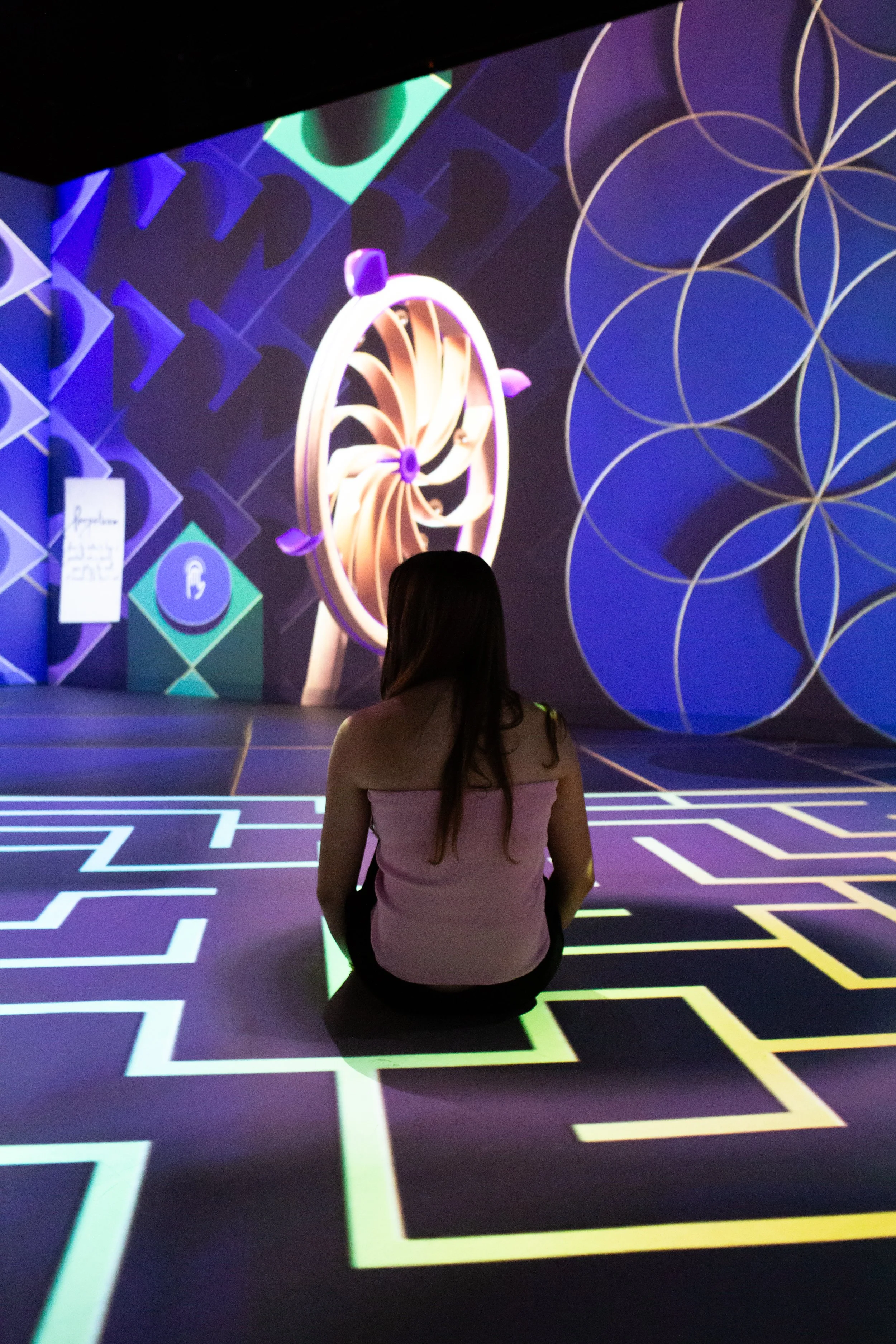 A woman sitting on a glowing green cube in a dark room with illuminated geometric patterns and digital art animations on the walls.