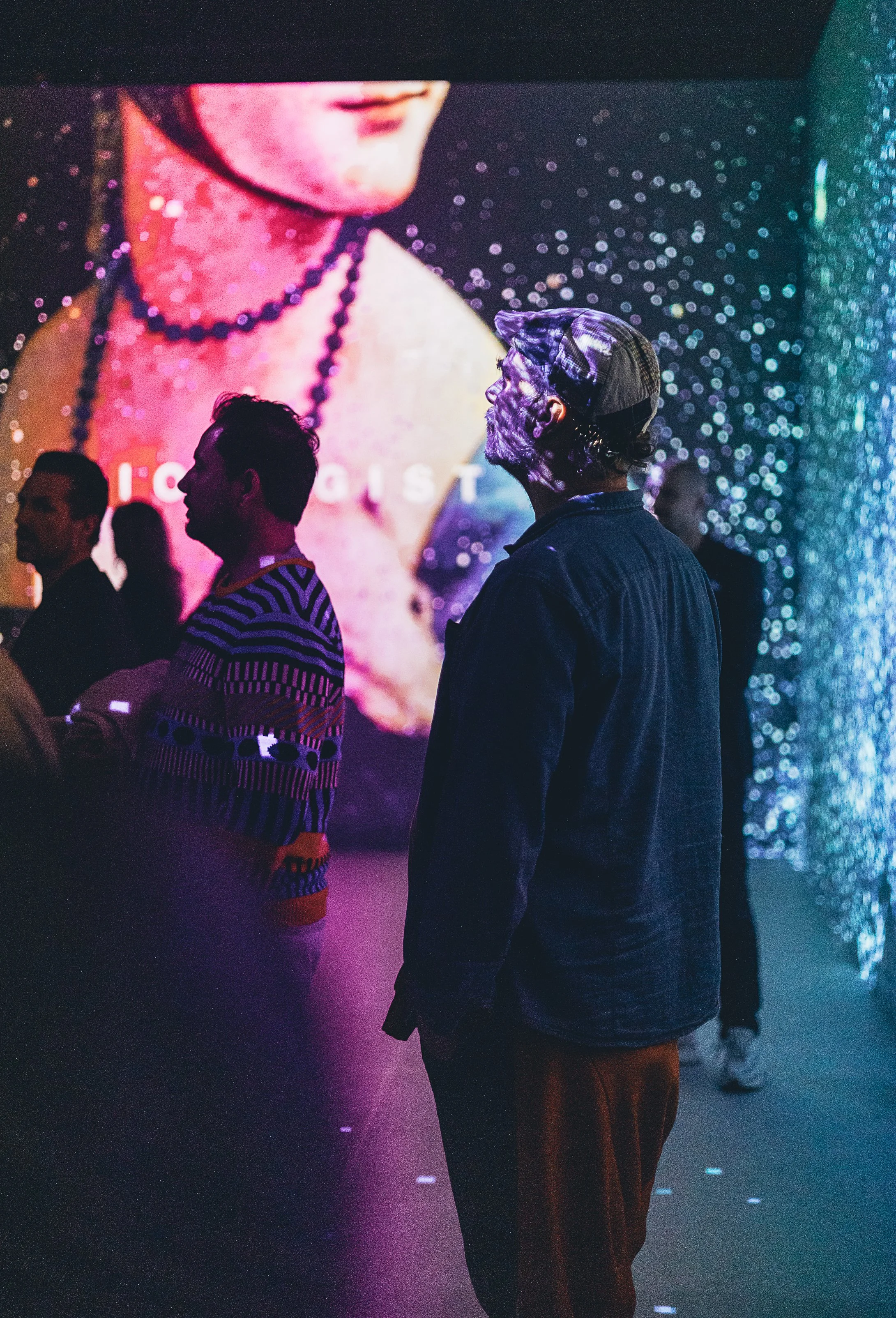People standing in a dimly lit space with a large digital screen displaying a woman's face in the background. The scene has colorful, vibrant lighting.