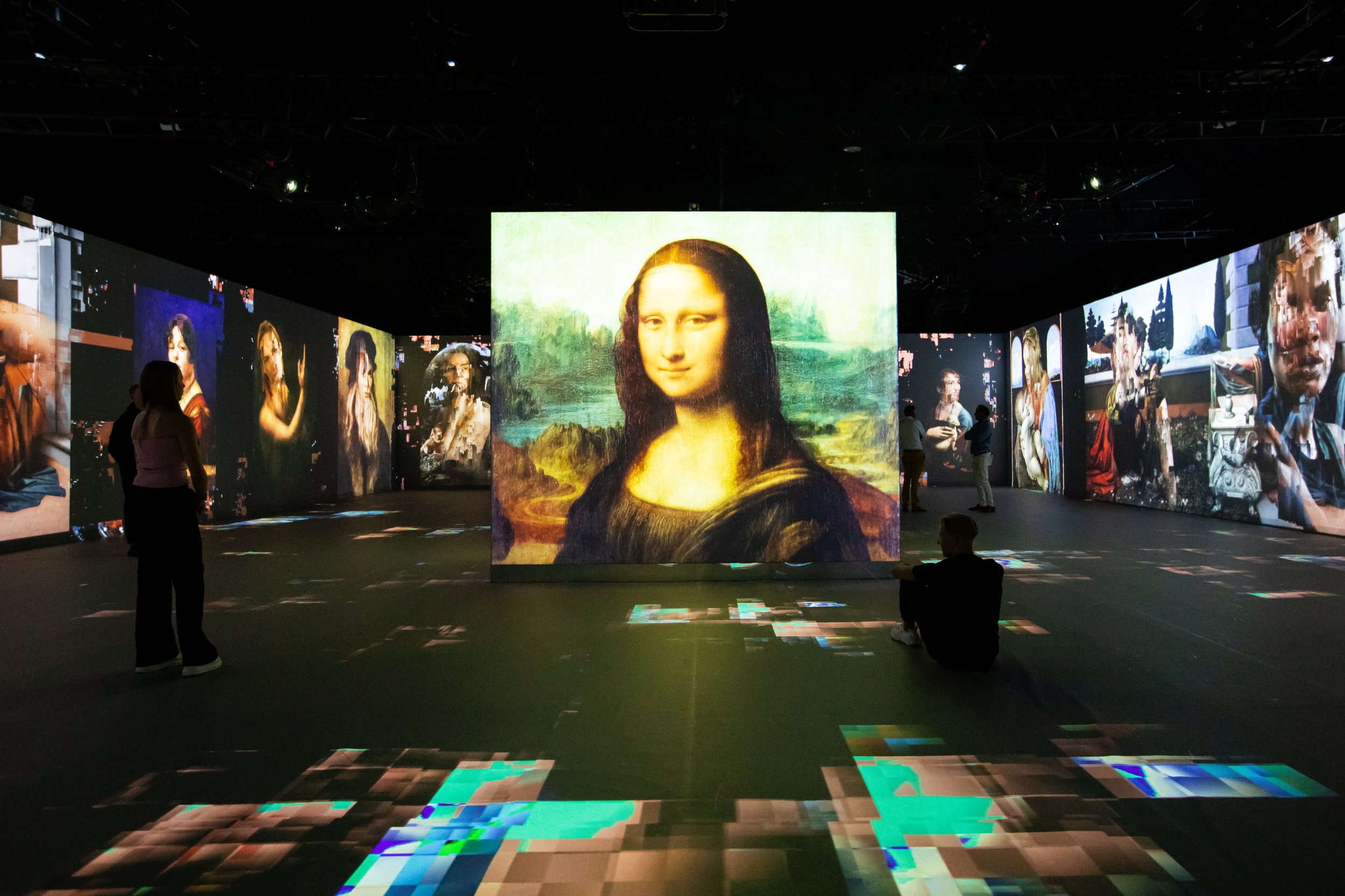 An immersive digital art exhibit featuring a large projection of the Mona Lisa surrounded by smaller digital images of her in various artistic styles and settings, with visitors observing the display.