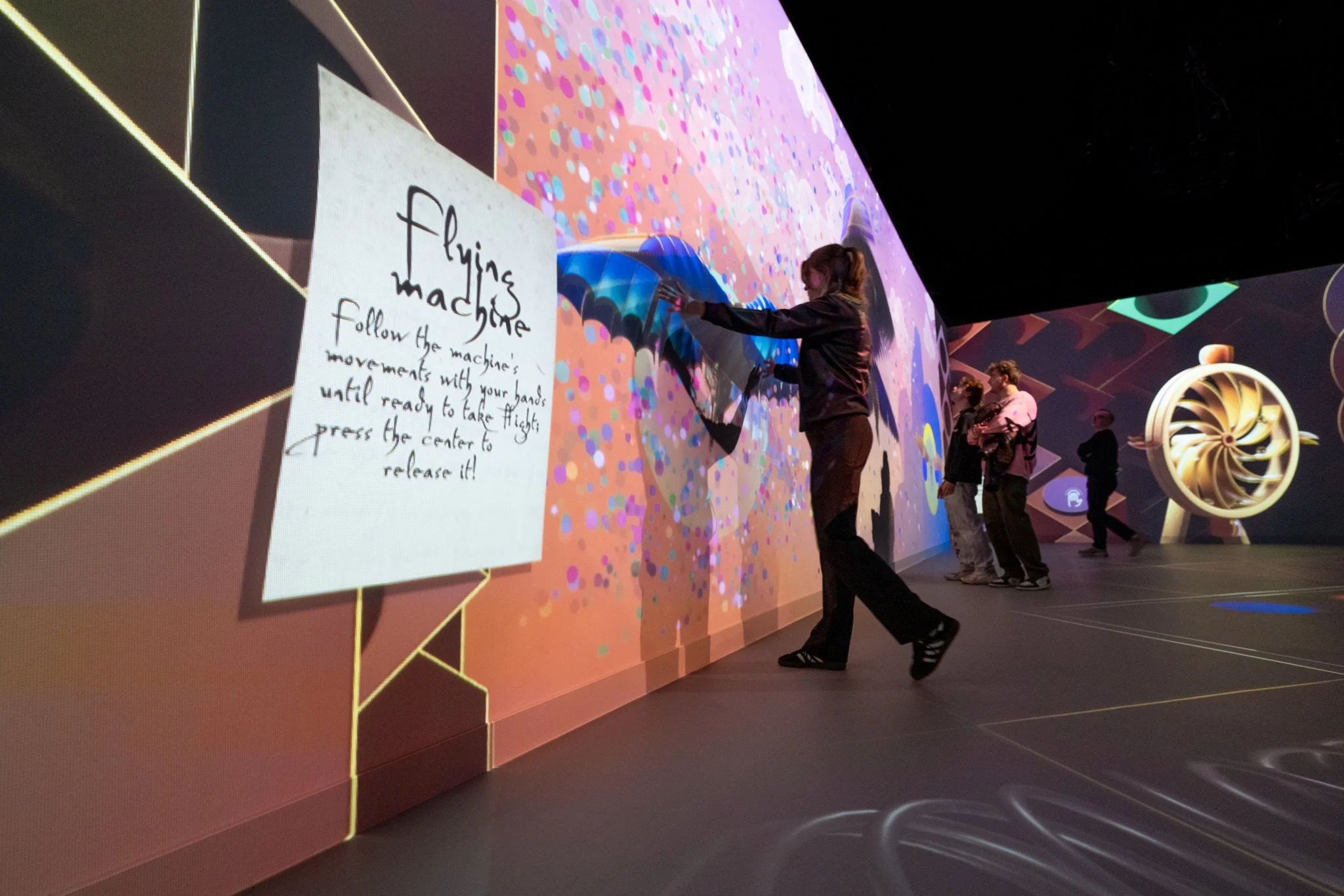 People interacting with an immersive digital art exhibit featuring a wall with colorful, animated visuals and a sign that reads 'flying machine' with instructions to follow the machine's movements with hands until ready to take flight.