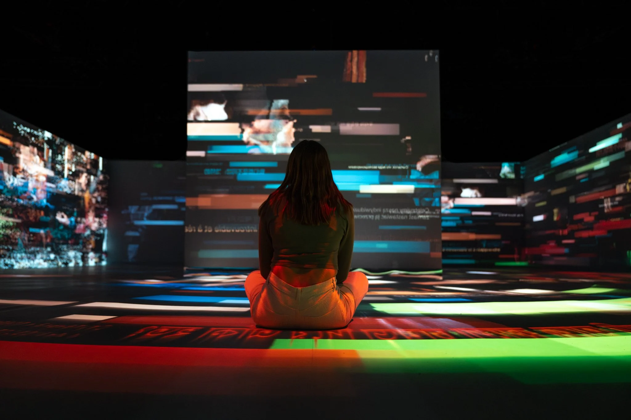 A person sitting cross-legged on the floor in a dark room, facing a large digital screen with colorful, distorted graphics and text.