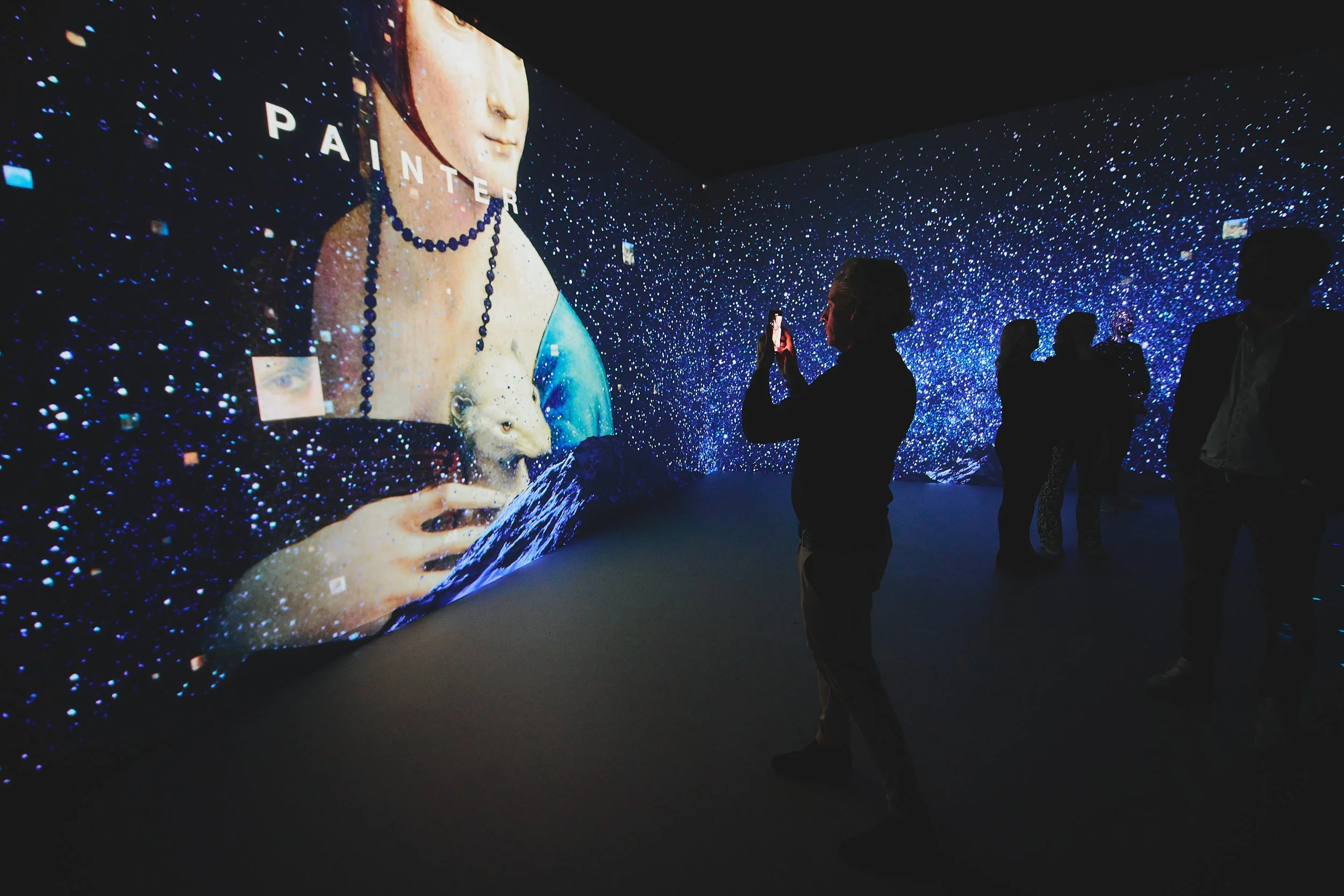 People in a dark room viewing a large digital art display showing the Mona Lisa with a polar bear coming out of her chest, surrounded by a starry background.