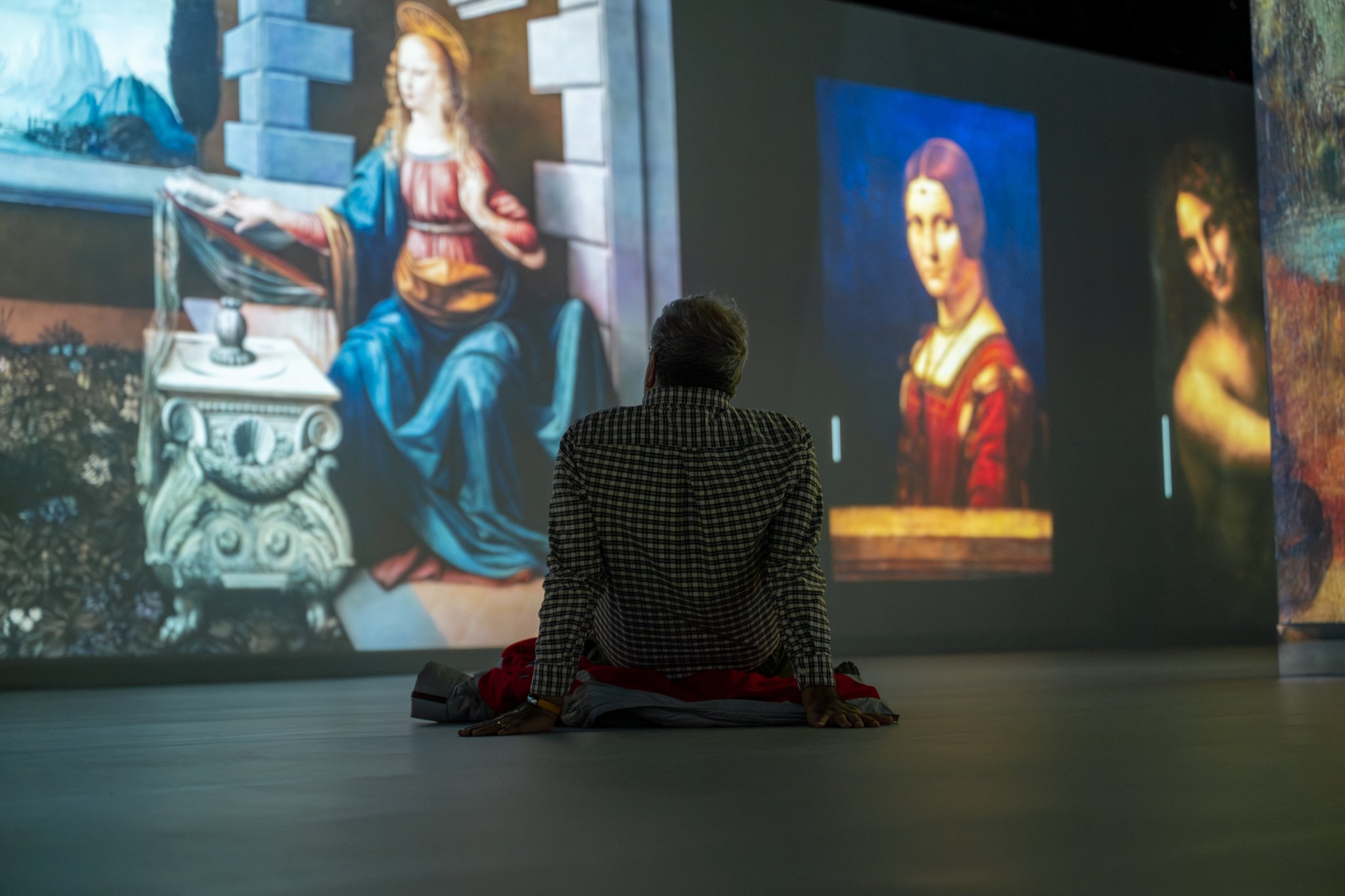A person sitting on the floor in an art gallery, viewing large digital screens displaying famous paintings, including the Mona Lisa and The Last Supper.