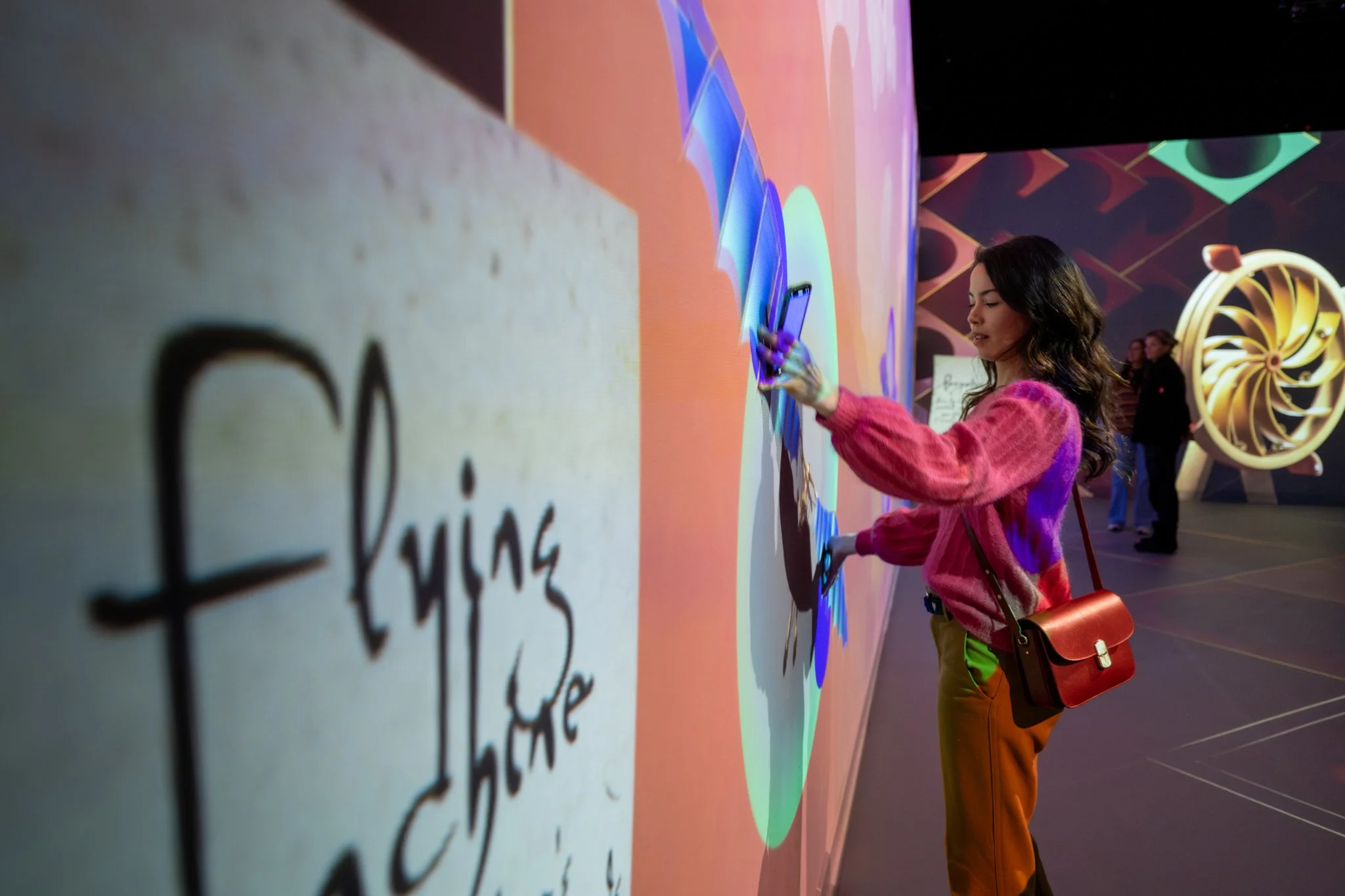 A woman with wavy dark hair interacts with an illuminated digital wall display at a modern exhibition or event, wearing a pink sweater and orange pants, carrying a small brown handbag.