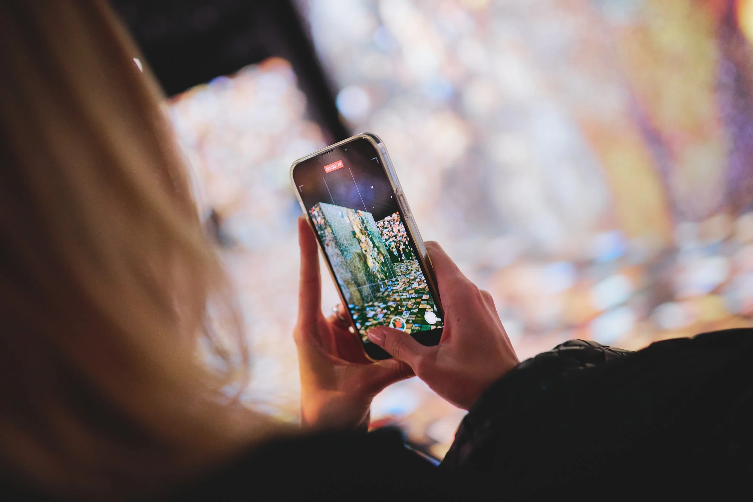 Person taking a photo with a smartphone in front of a colorful, illuminated background.