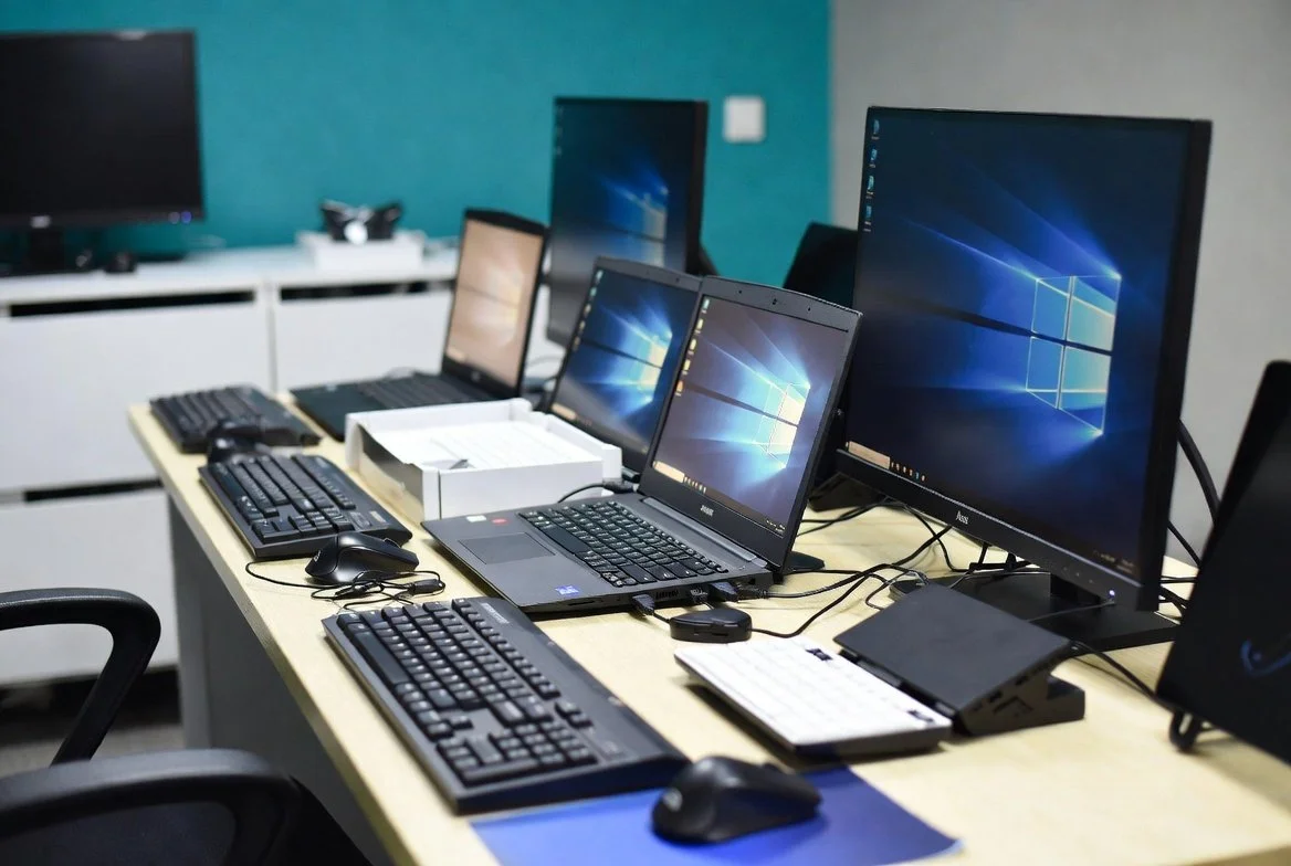 Office desk with multiple computer monitors, keyboards, mice, and laptops in a tech workspace.