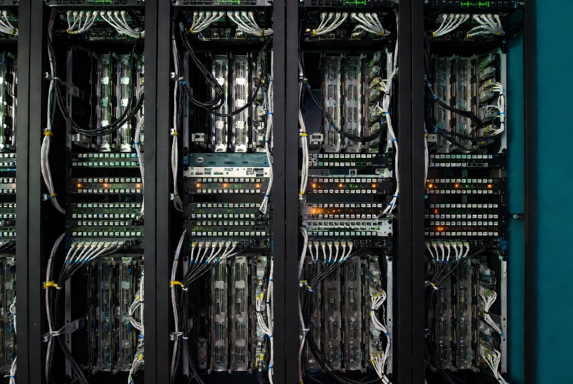 Interior of a data server room with multiple black server racks filled with computer hardware, cables, and blinking indicator lights.