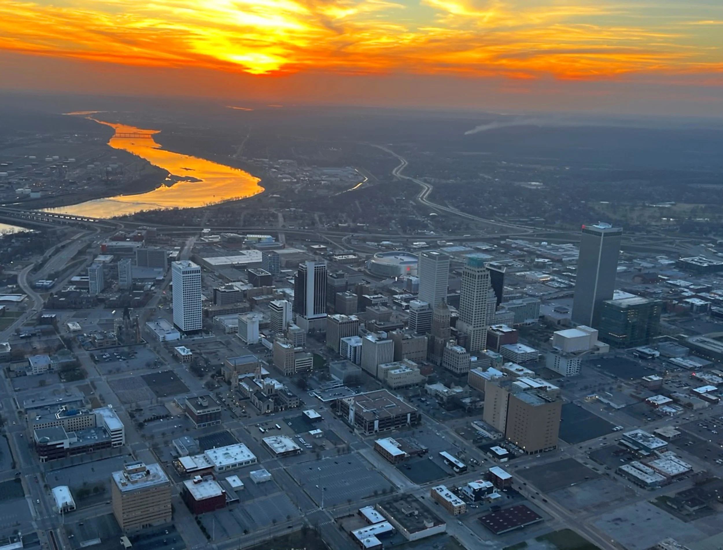 Aerial view of a city at sunset with a river reflecting the orange and yellow sky, surrounded by skyscrapers and roads.