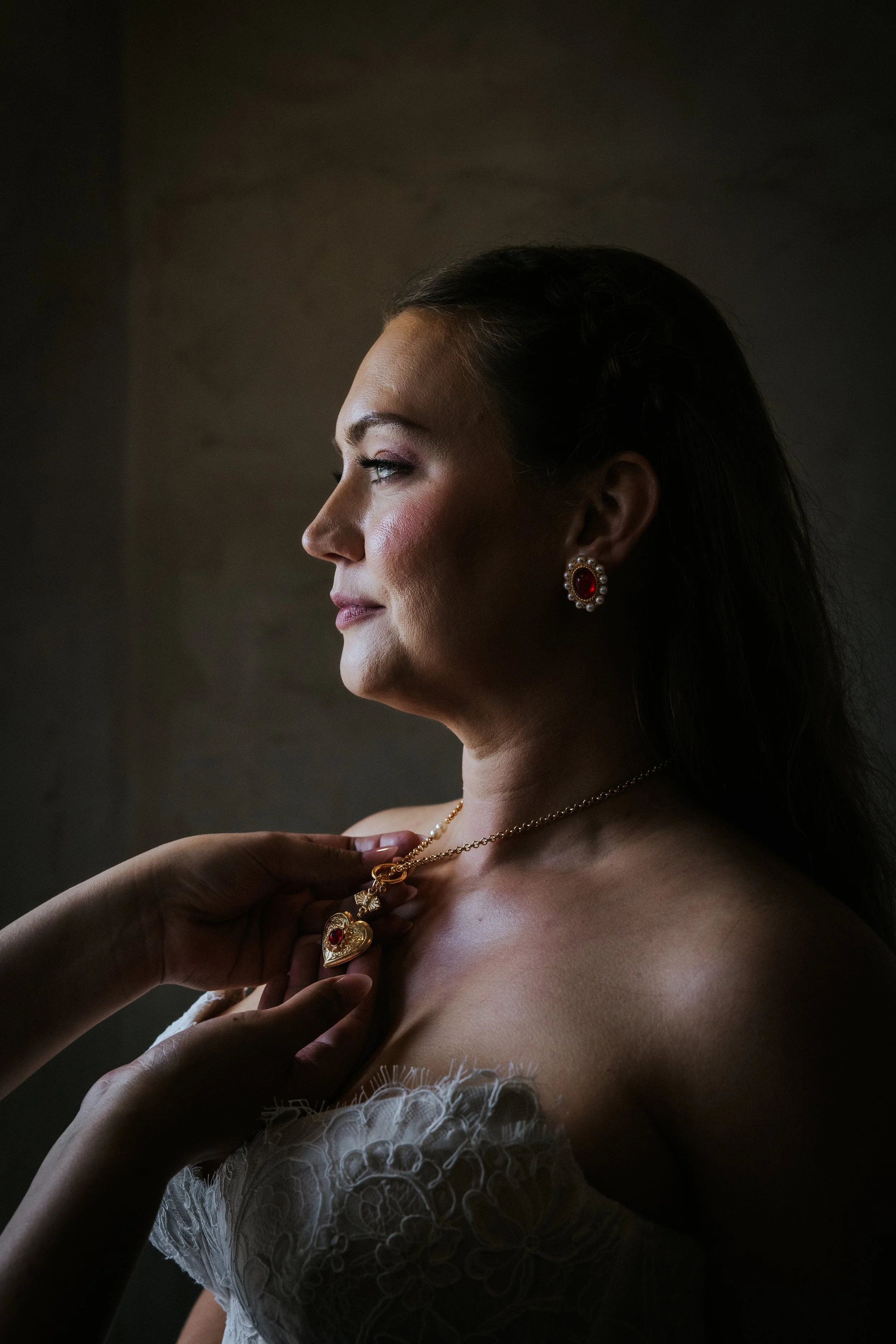 A woman in a lace wedding dress facing toward a window, with someone adjusting her gold heart necklace.