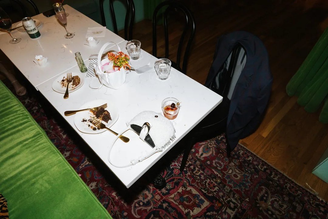 A white table with empty plates, glasses, a flower basket, and some leftover desserts. There are chairs around the table, some with jackets, and a green curtain on the side. The floor is wooden with a red patterned rug.