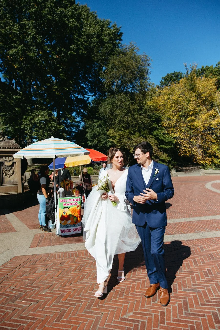Bride and groom walking outdoors on a brick path on a sunny day. The bride is in a white wedding dress holding a bouquet, and the groom is in a blue suit with brown shoes. There are trees, colorful umbrellas, and a small stall selling mangoes in the background.