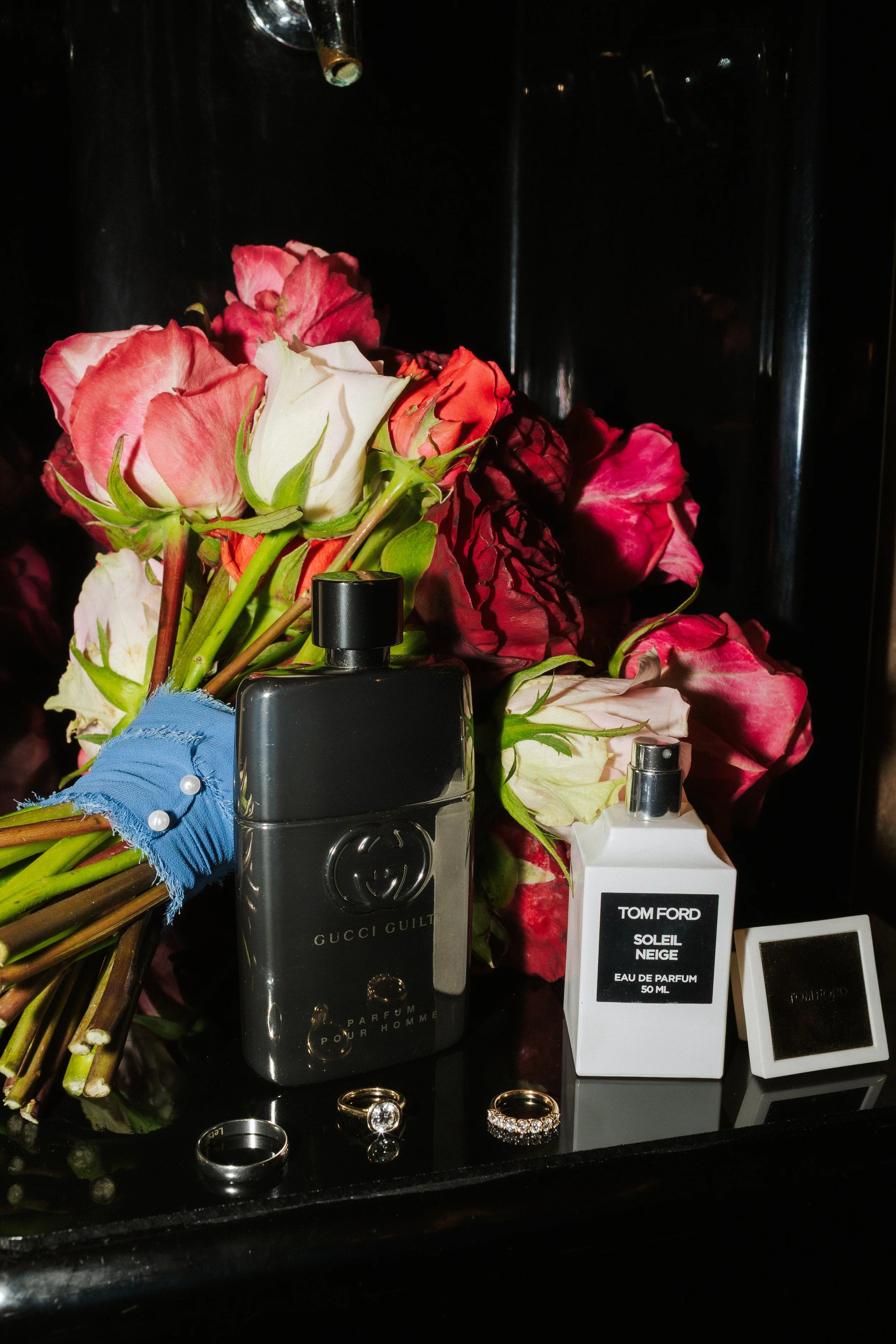 A bouquet of pink, white, and red roses, a pair of gold rings, and a few bottles of perfume on a black surface.