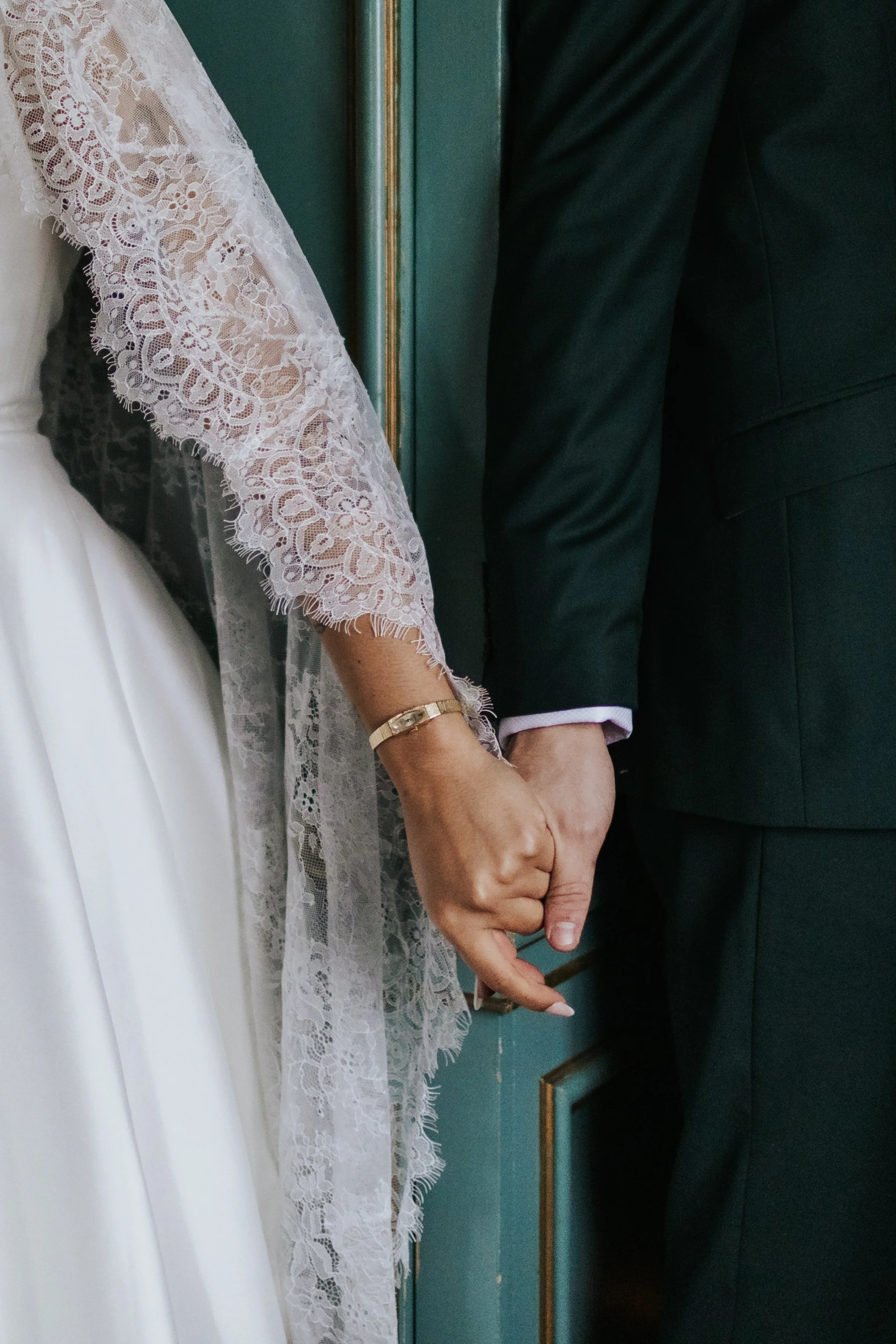 A bride and groom holding hands, with the bride wearing a lace sleeve wedding dress and gold bracelet, and the groom in a black suit, standing against a green door.